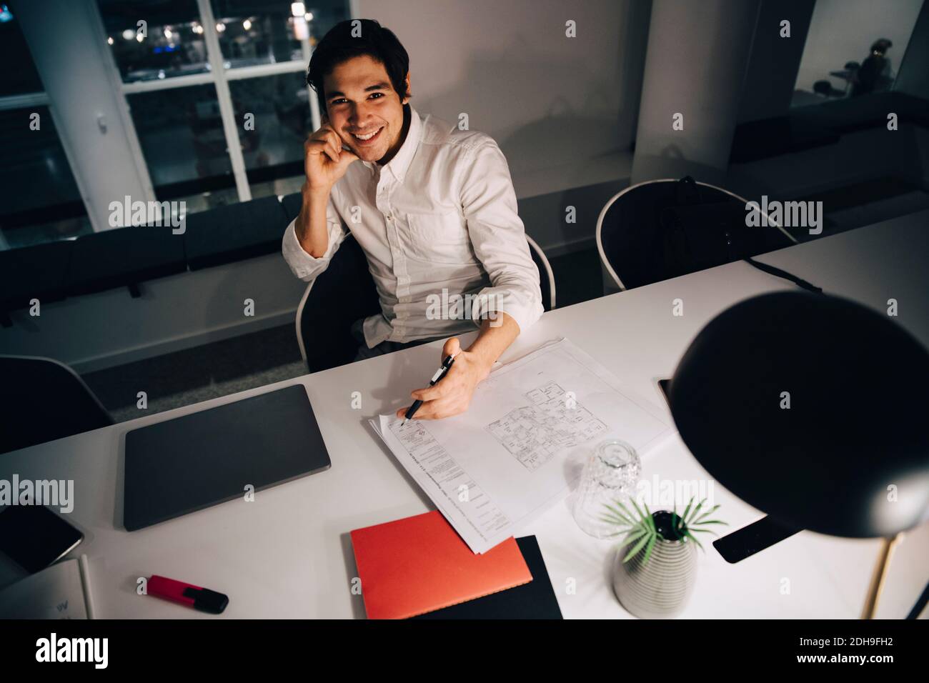 Portrait en grand angle d'un homme d'affaires souriant assis avec un plan d'action à bureau en travaillant tard dans le bureau Banque D'Images