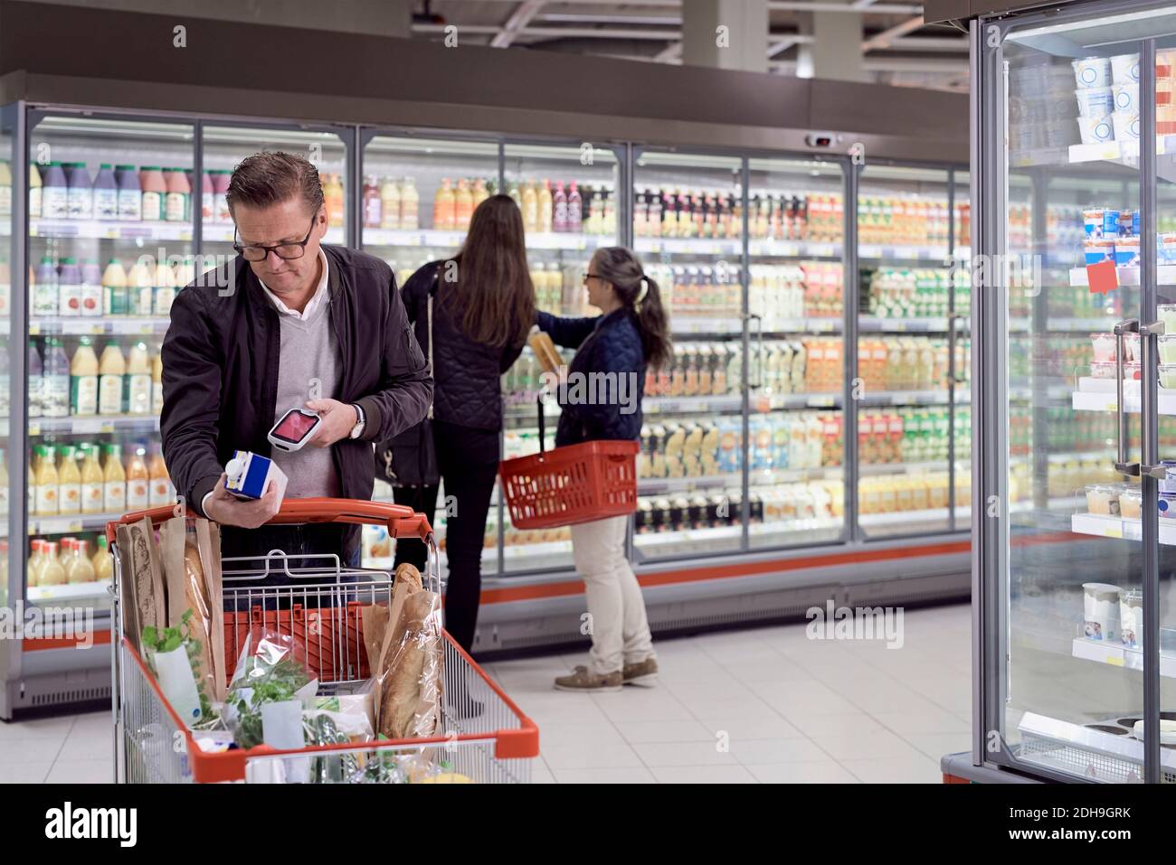 Homme mûr scannant boîte de jus avec lecteur contre les femmes achetant à la section réfrigérée dans le supermarché Banque D'Images