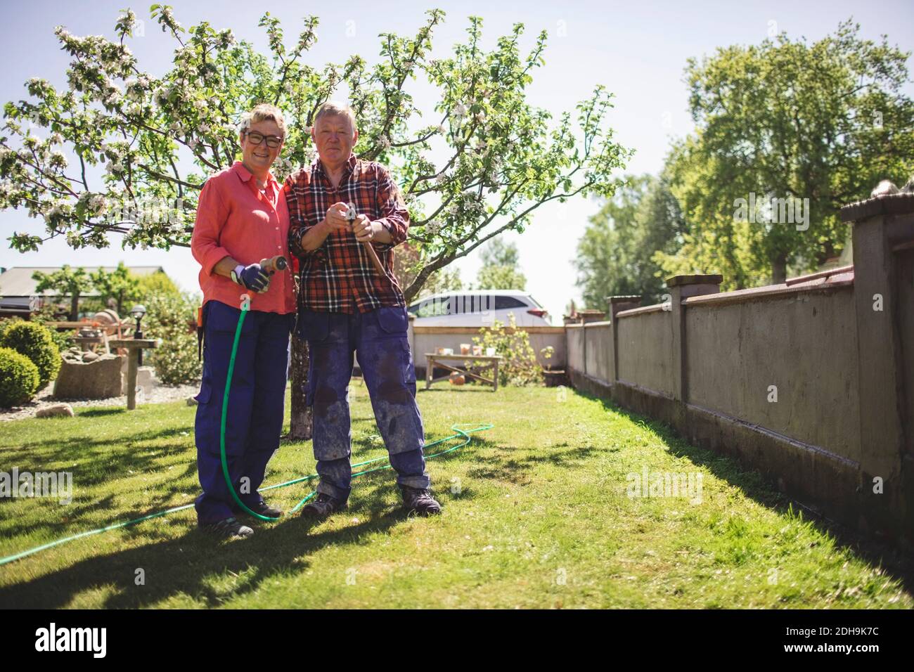 Portrait complet d'un couple senior tenant un marteau et un jardin tuyau en vous tenant à la cour Banque D'Images Portrait complet d'un couple senior tenant un marteau et un jardin tuyau en vous tenant à la cour Banque D'Images