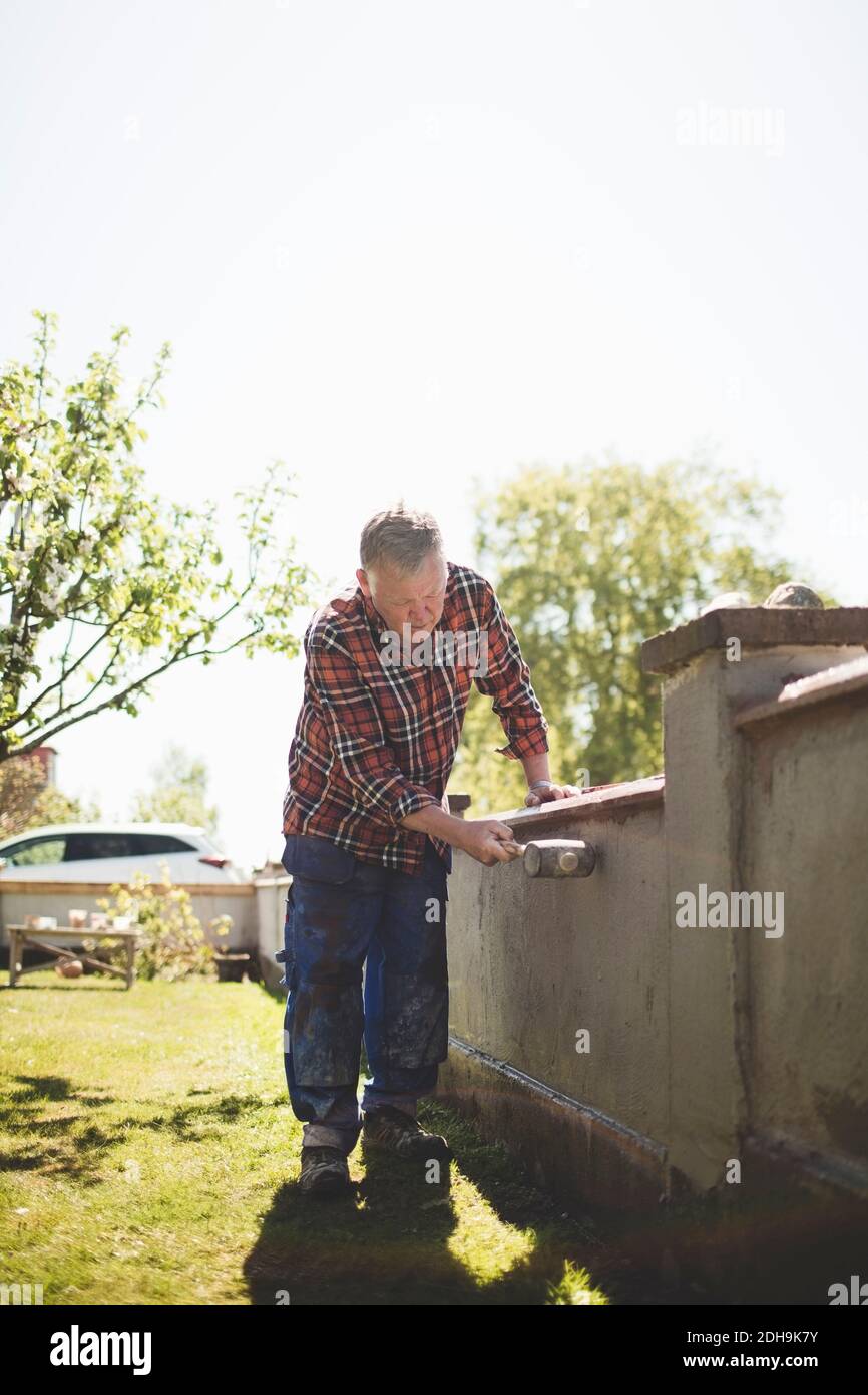 Pleine longueur d'homme senior examinant un mur en béton avec un marteau à la cour contre le ciel clair Banque D'Images Pleine longueur d'homme senior examinant un mur en béton avec un marteau à la cour contre le ciel clair Banque D'Images
