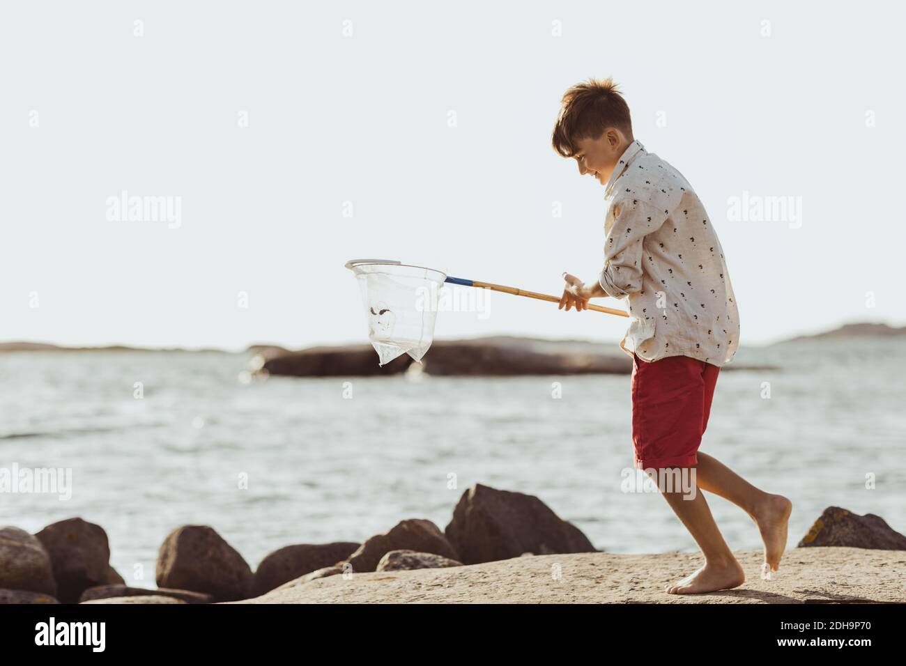 Garçon souriant avec filet de pêche marchant sur la formation de roche contre ciel dégagé pendant la journée ensoleillée Banque D'Images
