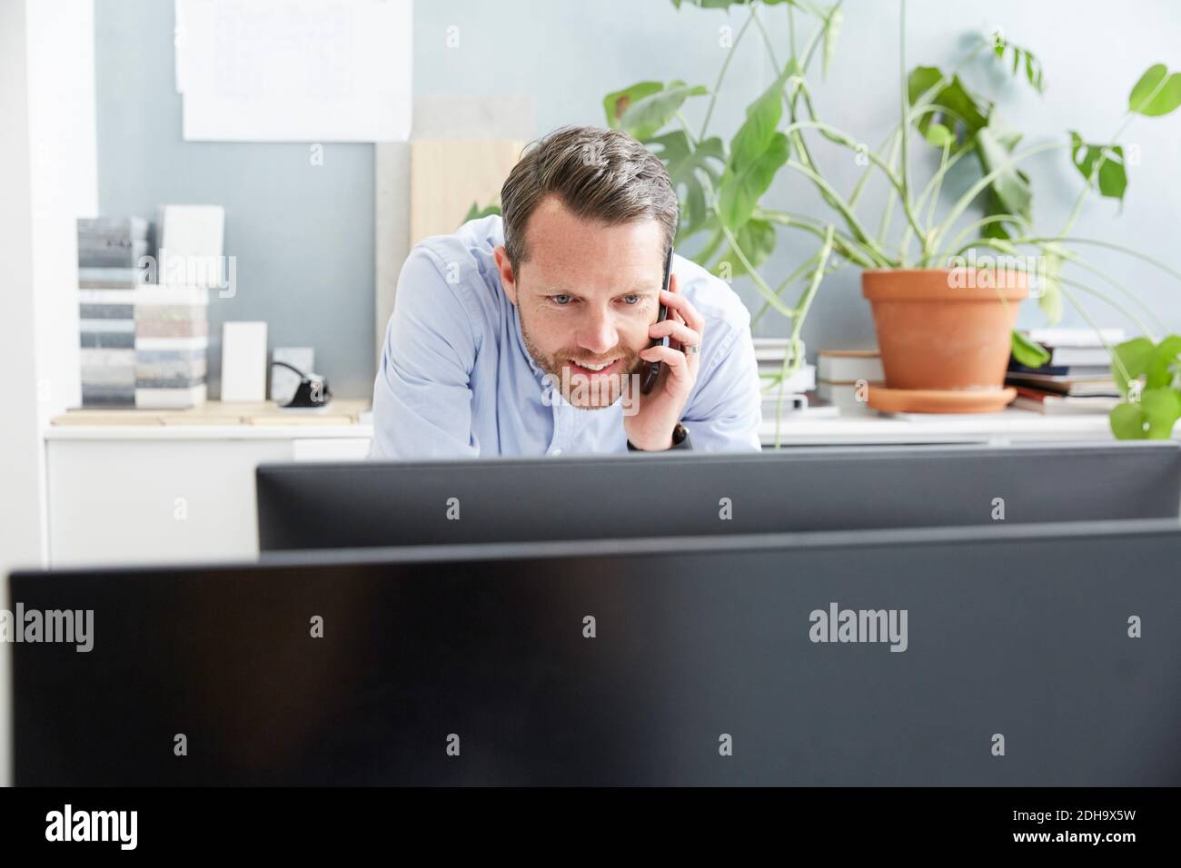 Homme d'affaires souriant parlant sur un téléphone portable tout en utilisant l'ordinateur à bureau créatif Banque D'Images