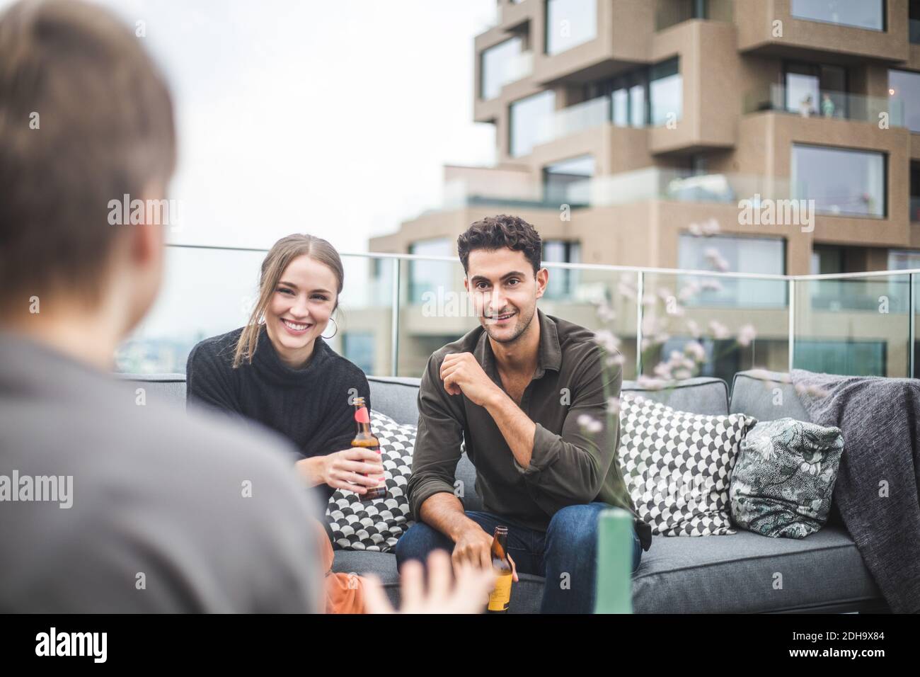 Homme souriant et femme regardant une amie pendant qu'elle est assise sur le canapé pendant la fête sur la terrasse du bâtiment Banque D'Images