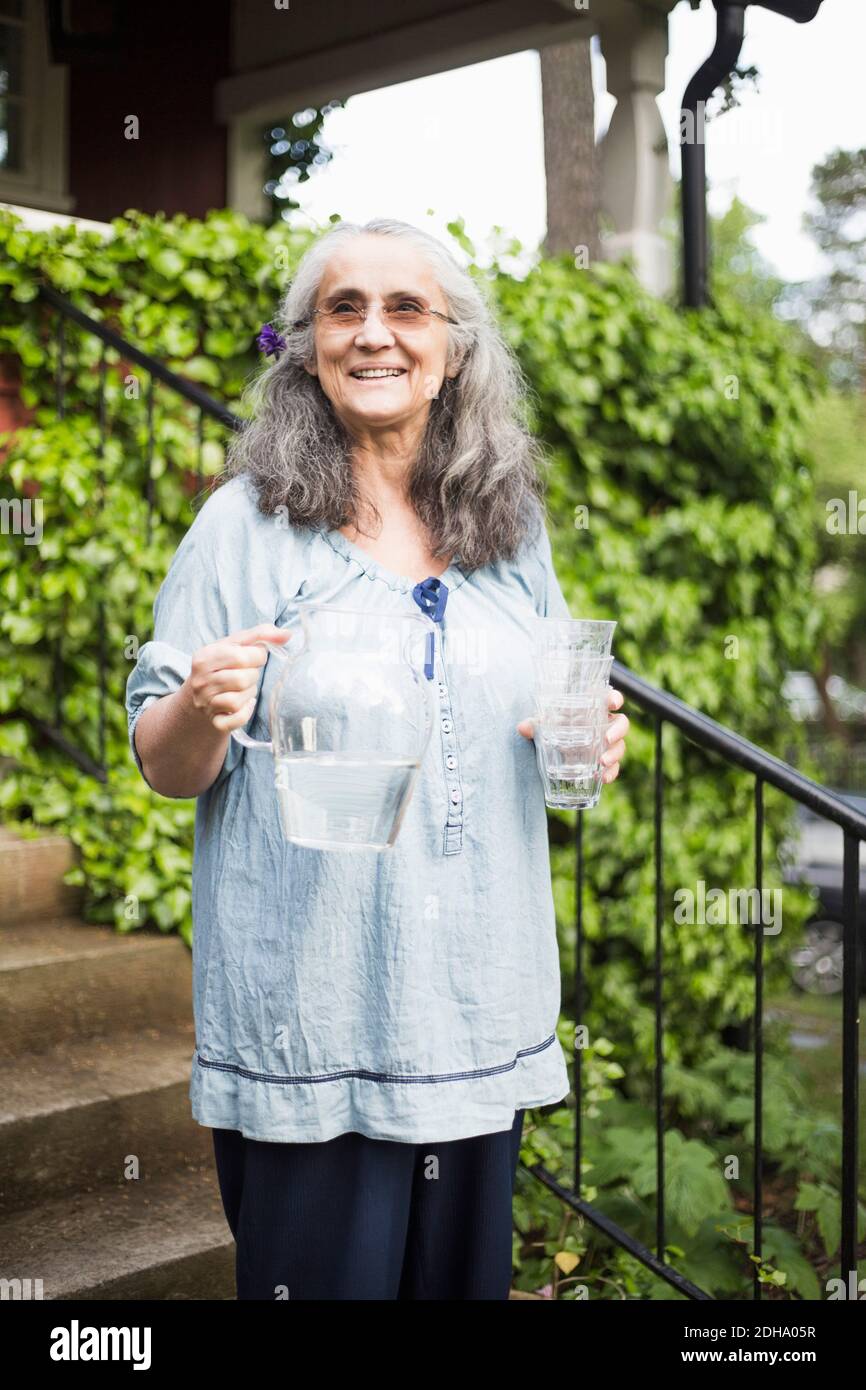 Femme âgée souriante debout avec pichet à eau et verre dedans arrière-cour Banque D'Images