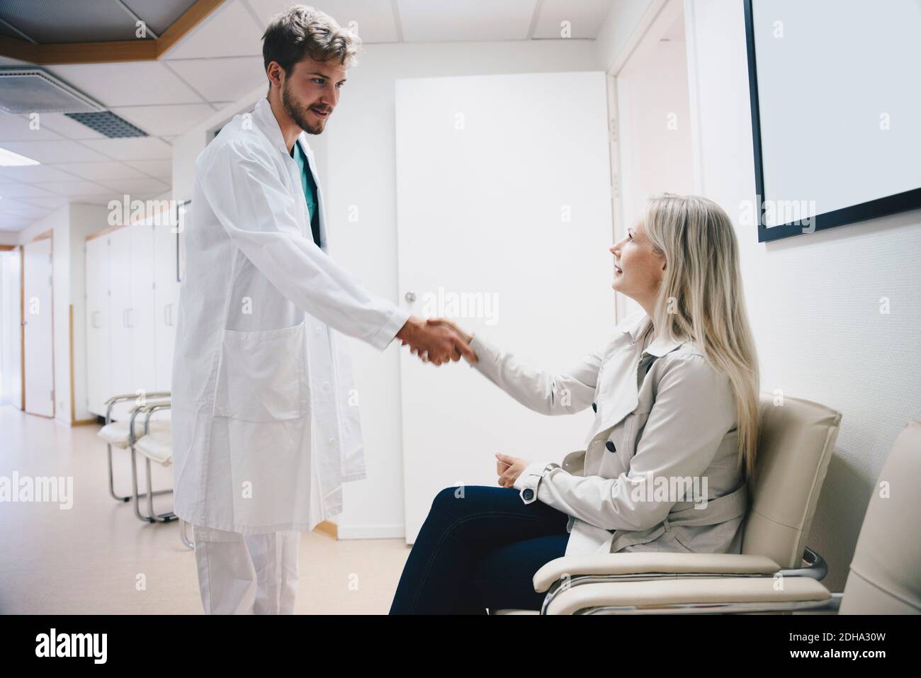 Jeune homme médecin saluant femme patiente assise sur une chaise couloir de l'hôpital Banque D'Images
