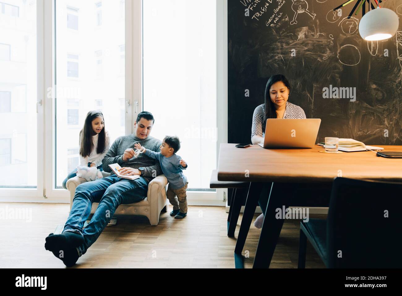 Mère utilisant un ordinateur portable à la table pendant que le père joue avec des enfants contre la fenêtre à la maison Banque D'Images