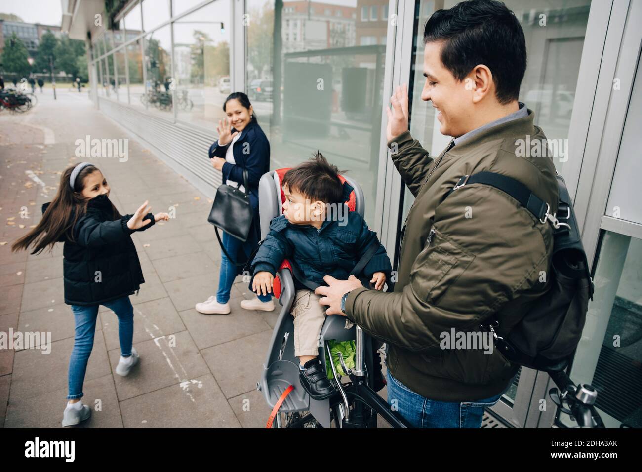 Une mère et une fille souriantes agitant un garçon avec son père trottoir en ville Banque D'Images