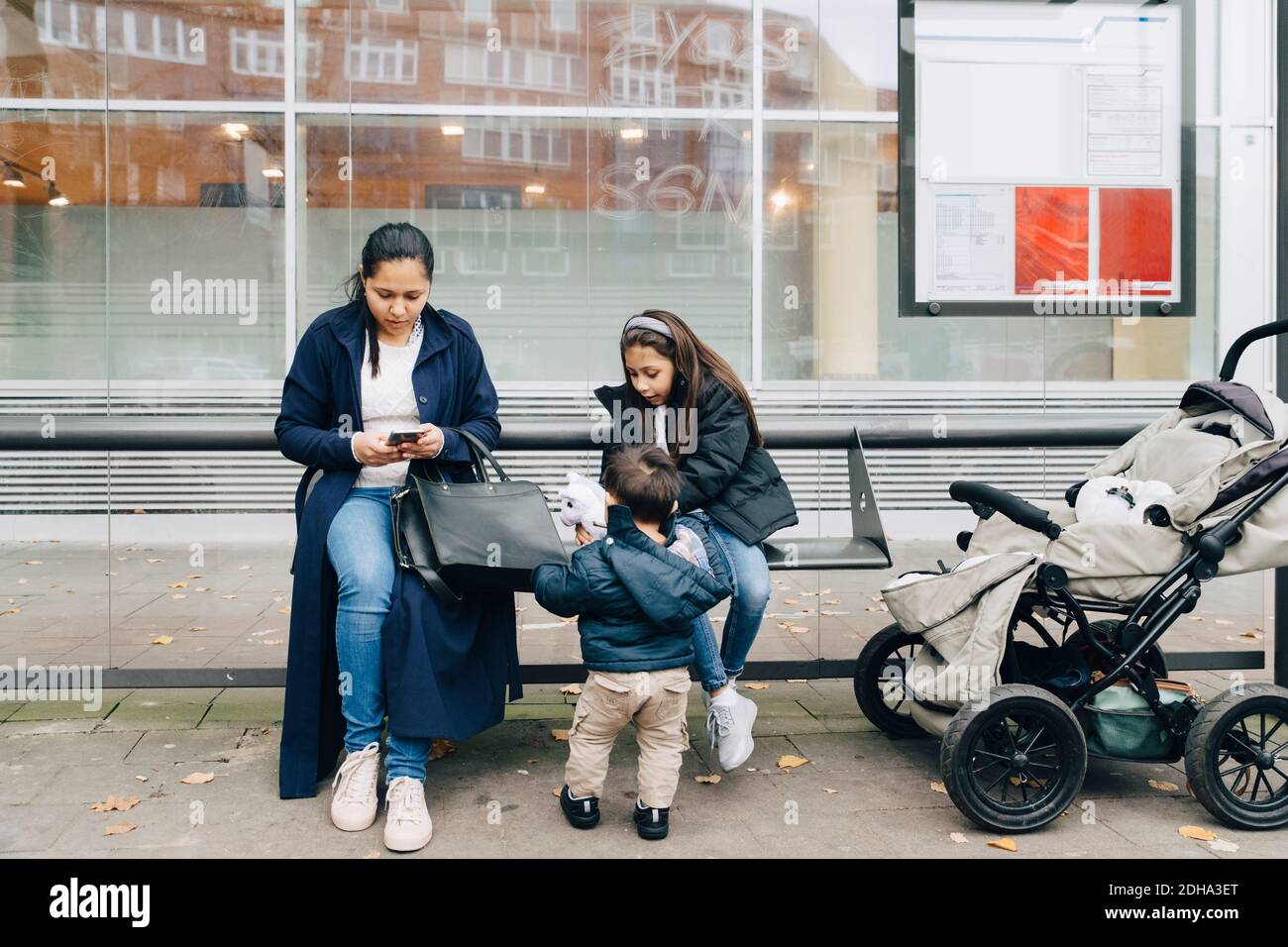 Mère utilisant un smartphone tout en étant assise avec des enfants dans le bus arrêt en ville Banque D'Images