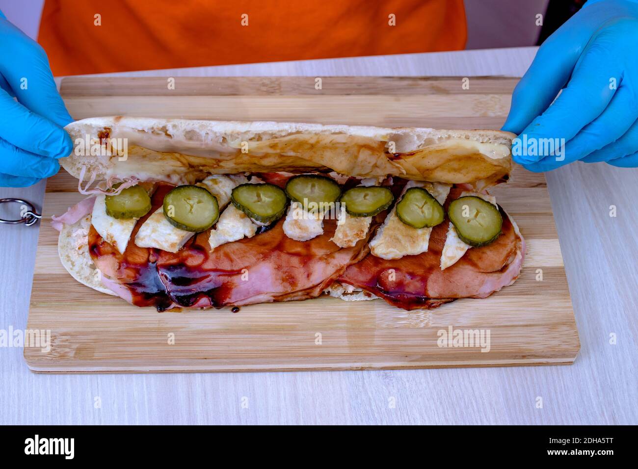 Chef préparant un délicieux sandwich sous-marin à la baguette fraîche. Gros plan des mains dans des gants fermant la baguette sur les tranches de cornichon. Banque D'Images