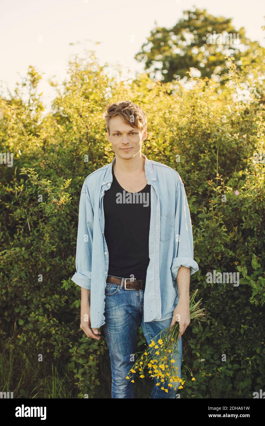 Portrait d'un jeune homme tenant des fleurs debout contre des plantes Banque D'Images