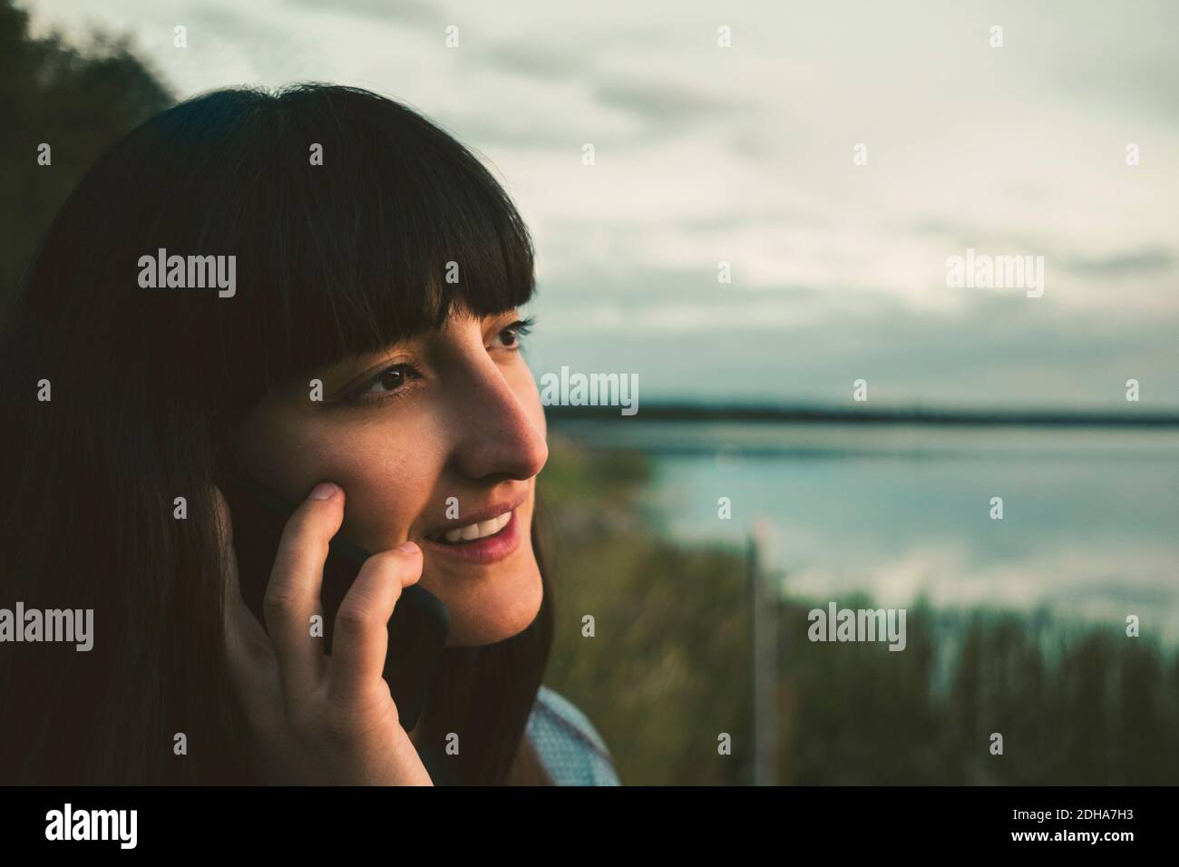 Femme souriante parlant sur téléphone portable près du lac contre le ciel Banque D'Images