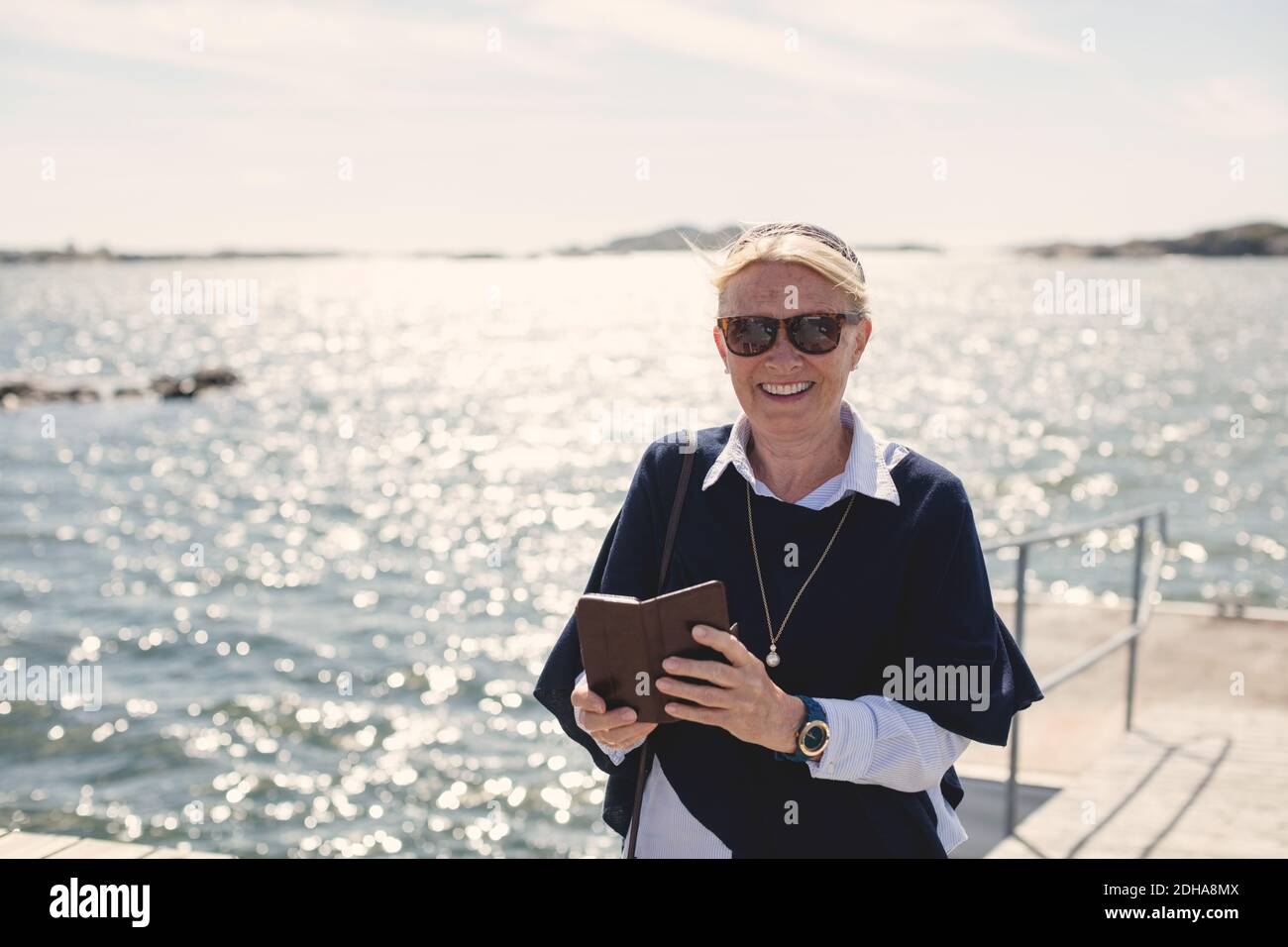 Portrait d'une femme âgée souriante portant des lunettes de soleil tout en tenant un mobile téléphone contre le lac Banque D'Images