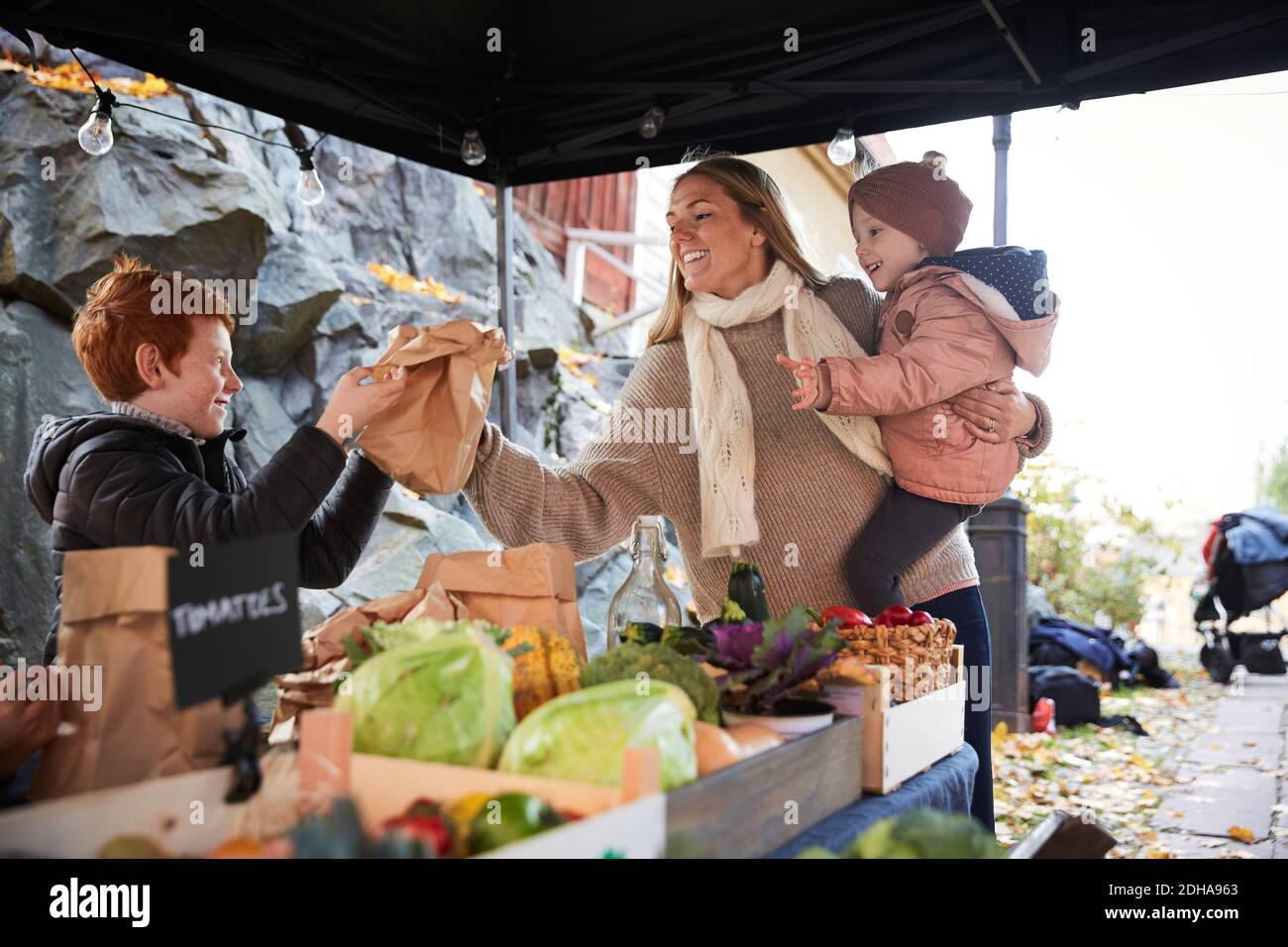 Femme souriante transportant sa fille et achetant des légumes auprès du vendeur du marché Banque D'Images