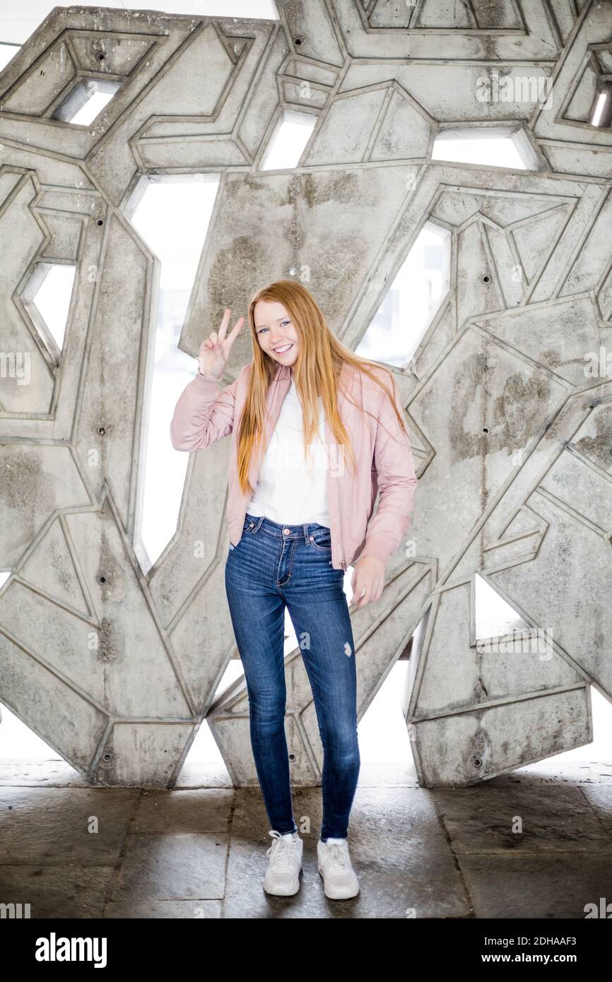 Portrait complet de fille blonde souriante montrant le signe de paix en vous tenant contre un mur en béton au garage Banque D'Images