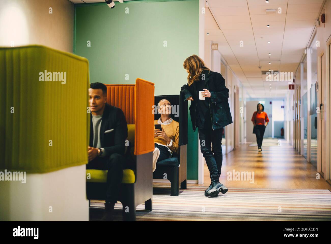 Pleine longueur de femme d'affaires debout dans le couloir avec un verre parlant à un collègue au bureau Banque D'Images