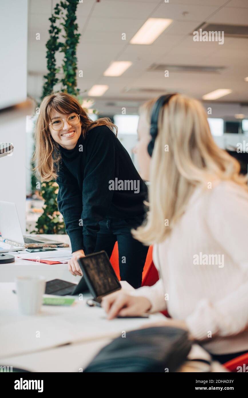 Une jeune femme d'affaires souriante s'est penchée sur son bureau tout en regardant une collègue féminine travailler au bureau Banque D'Images