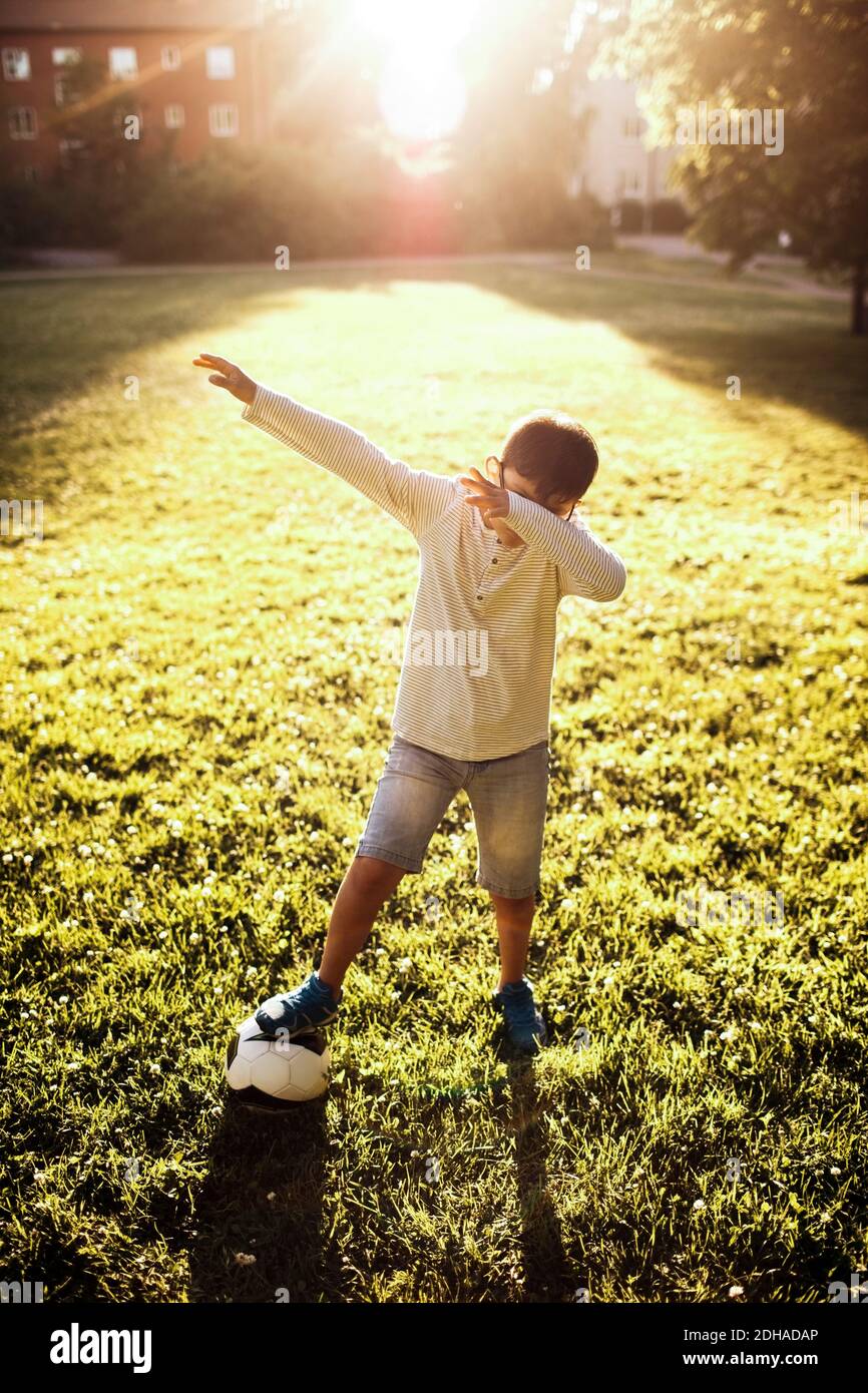 Longueur complète de garçon avec ballon de football couvrant le visage pendant se tenir sur un terrain herbeux au parc Banque D'Images