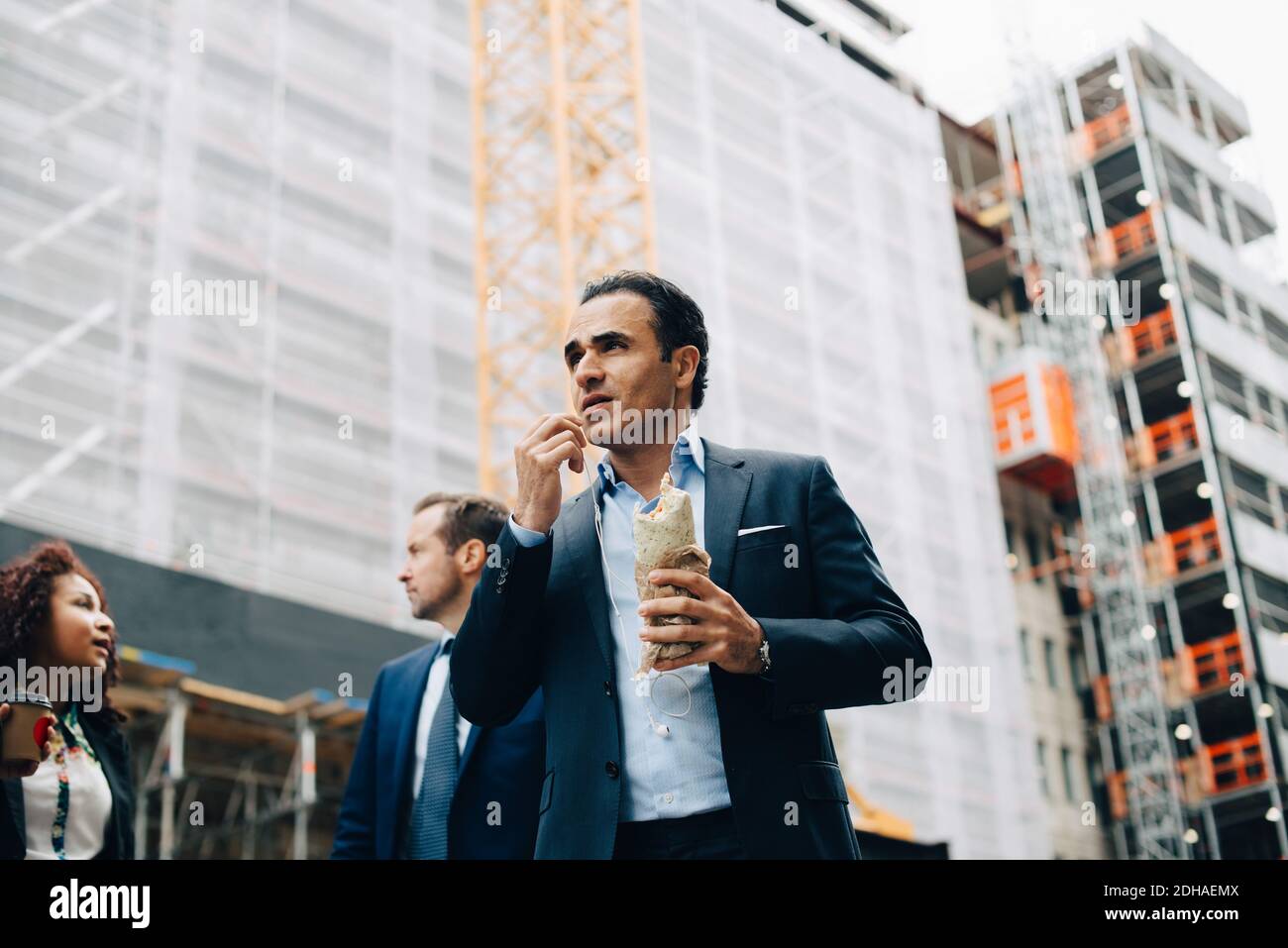 Vue à angle bas de l'homme d'affaires mangeant sandwich enveloppé tout en étant debout par des collègues contre la construction en ville Banque D'Images