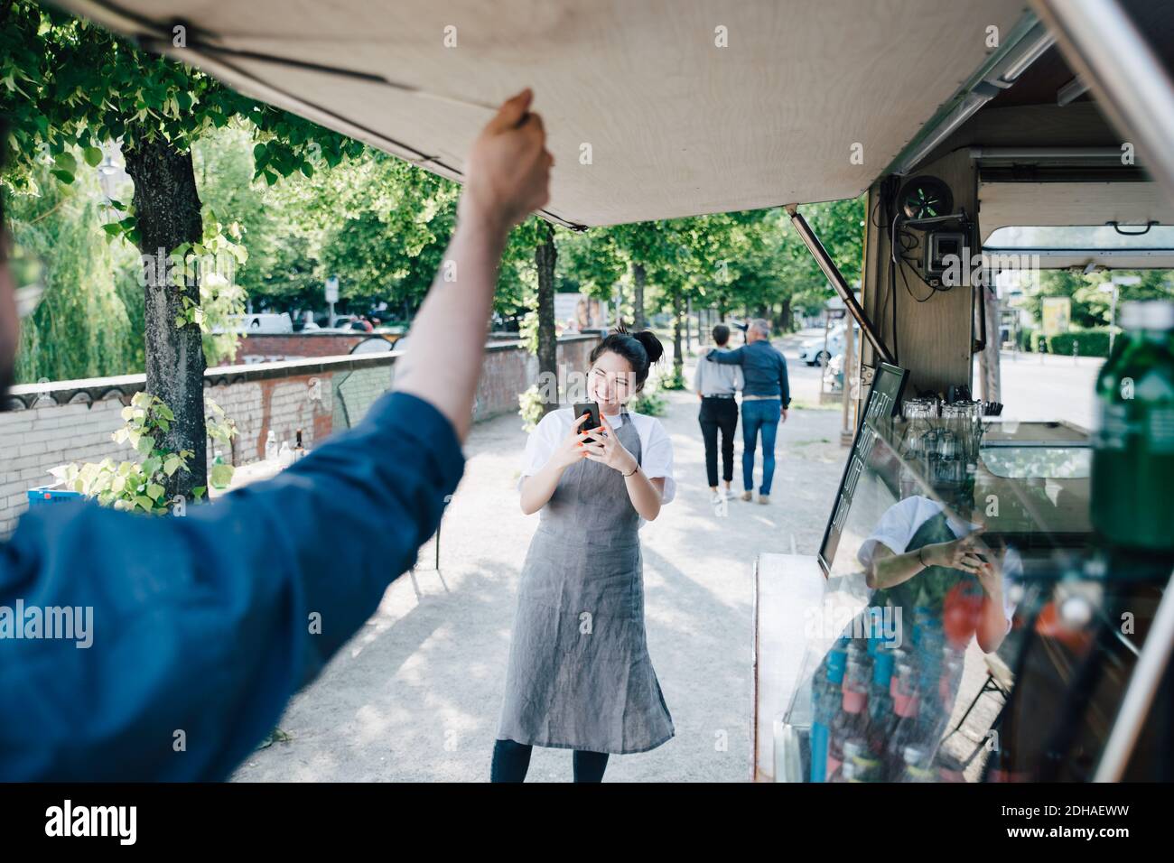 Femme propriétaire photographiant à l'aide d'un smartphone pendant que l'un de ses collègues ouvre abat-jour du camion alimentaire Banque D'Images