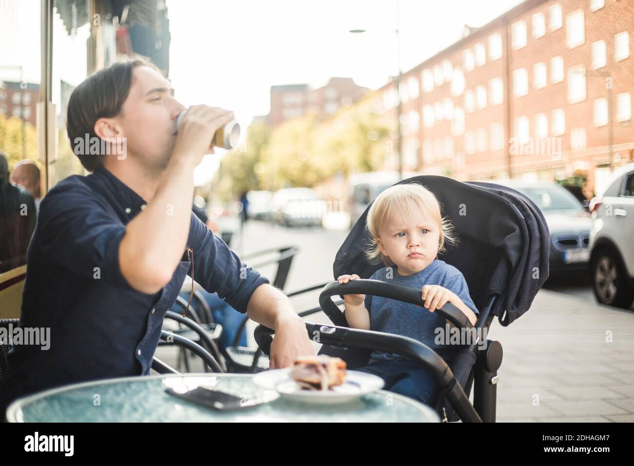 Bébé garçon regardant loin en étant assis sur la poussette par le père boire au café-terrasse de la ville Banque D'Images