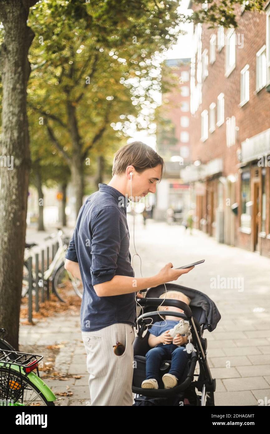 Vue latérale de l'homme utilisant un smartphone en étant debout fils sur poussette de bébé dans la ville Banque D'Images