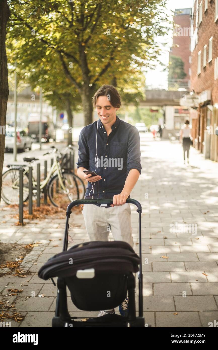 Homme souriant utilisant un smartphone tout en poussant la poussette de bébé sur sentier de la ville Banque D'Images