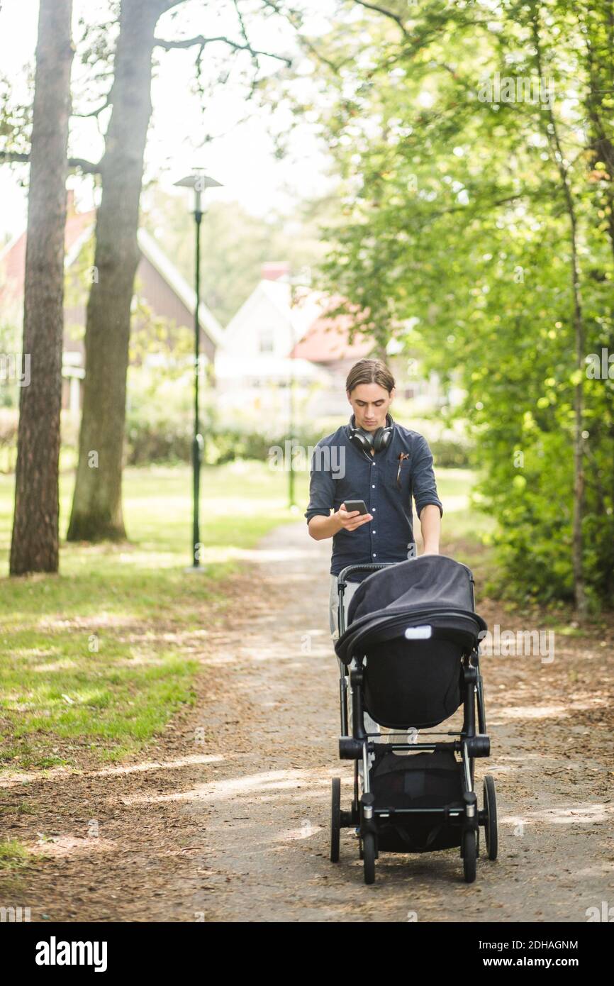 Homme utilisant un smartphone tout en poussant la poussette de bébé sur le chemin de pied au parc public Banque D'Images