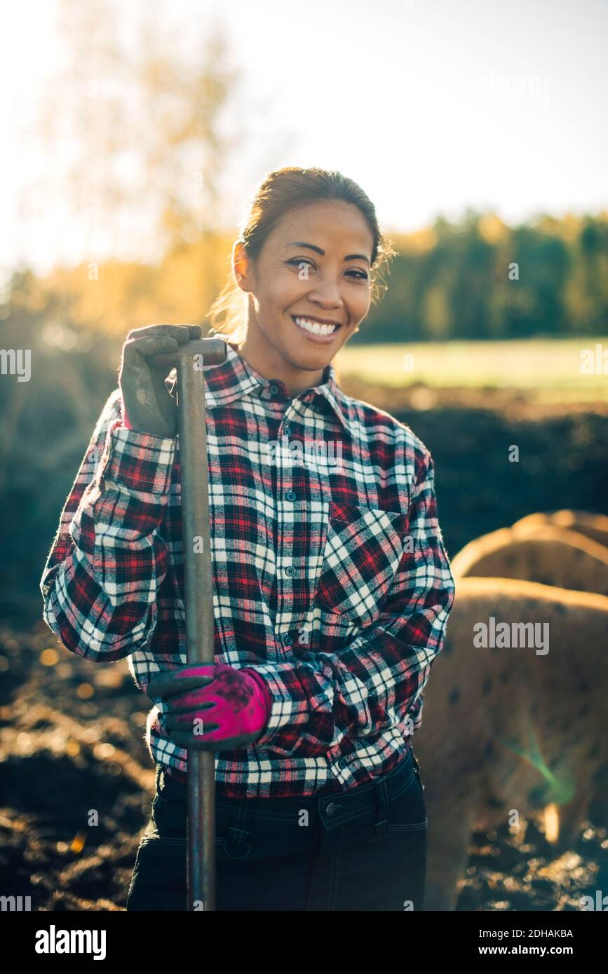 Portrait d'une agricultrice du milieu adulte souriante tenant une pelle avec porcs en arrière-plan à la ferme biologique Banque D'Images