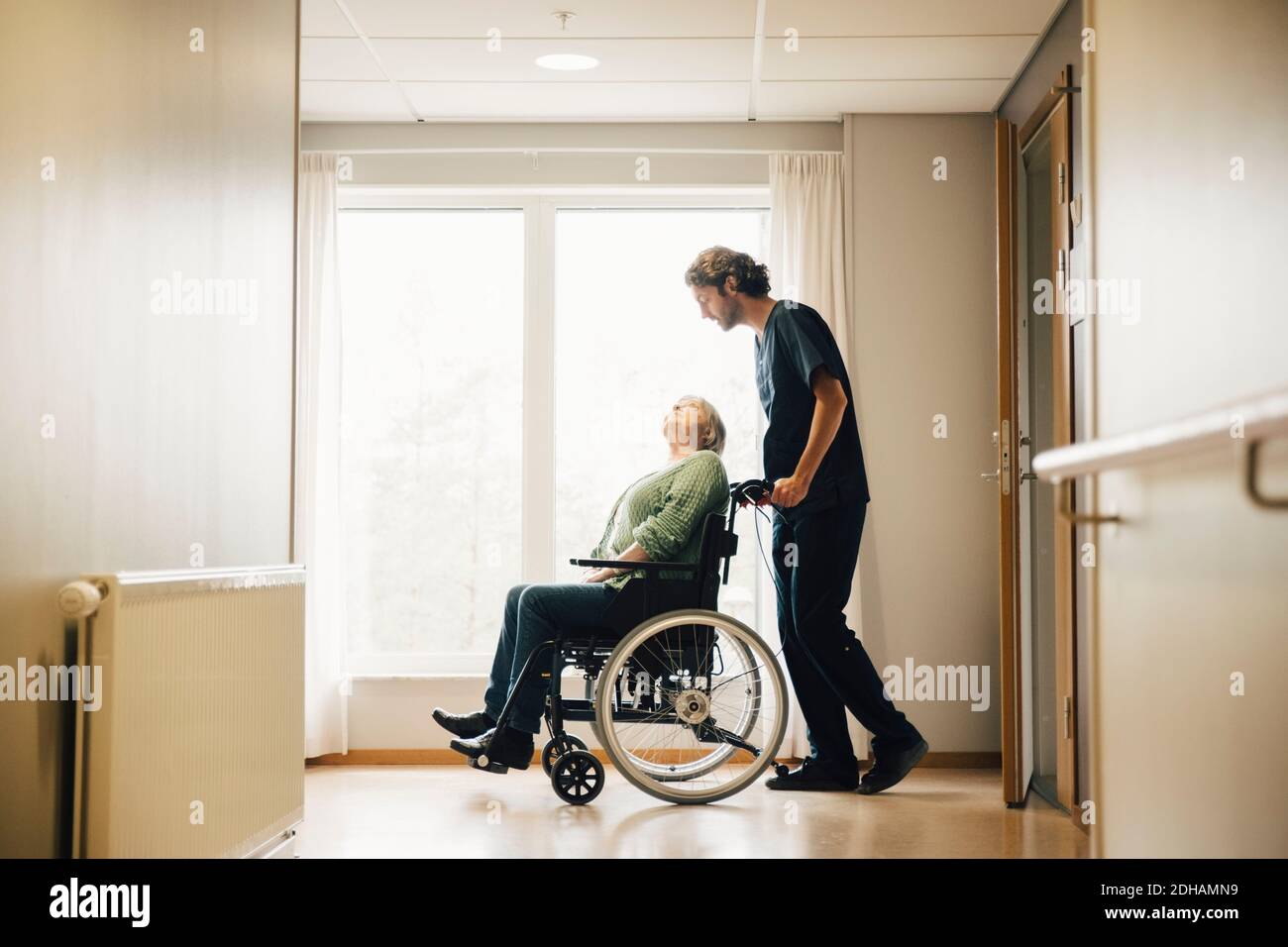 Vue latérale sur toute la longueur d'un infirmier poussant un aîné handicapé femme en fauteuil roulant dans une ruelle à la maison de retraite Banque D'Images