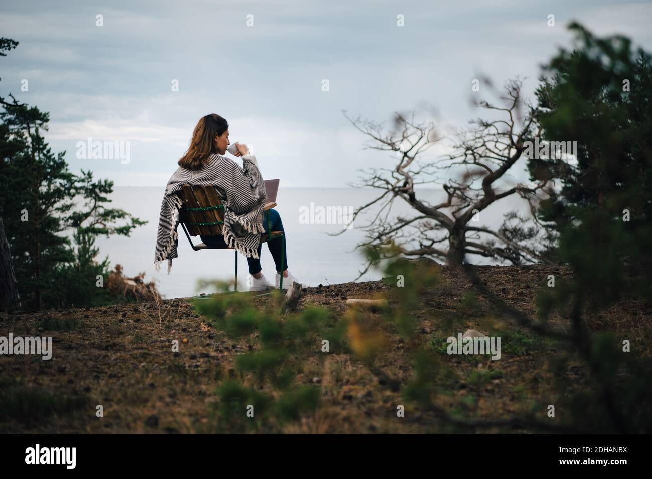 Vue arrière d'une femme buvant du café tout en étant assise avec un ordinateur portable sur chaise contre le lac Banque D'Images Vue arrière d'une femme buvant du café tout en étant assise avec un ordinateur portable sur chaise contre le lac Banque D'Images