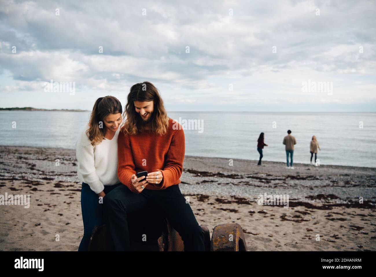 Couple regardant le téléphone portable en étant assis sur la mer à la plage Banque D'Images