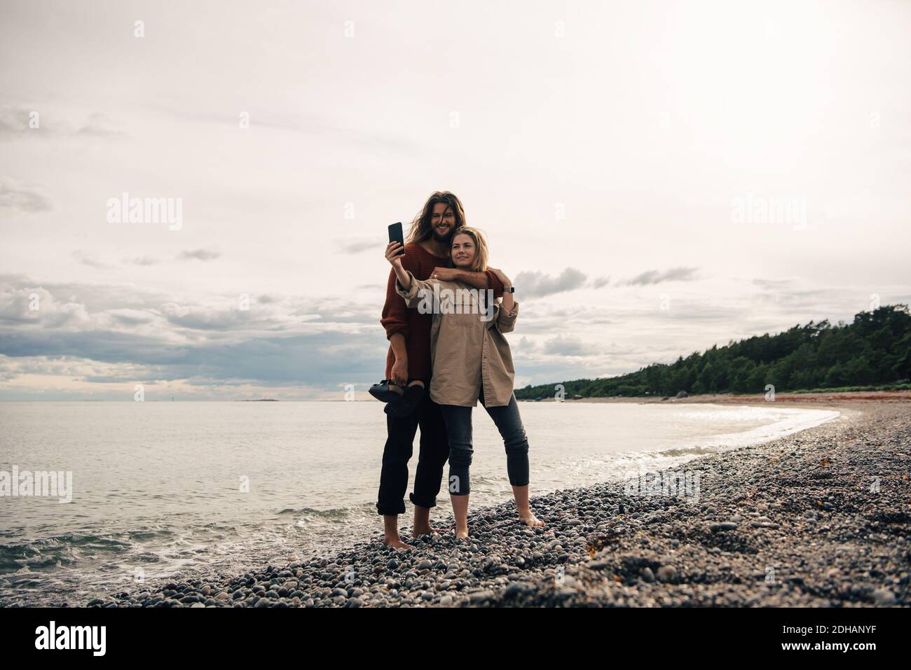 Couple heureux de prendre selfie sur smartphone à la plage contre ciel au coucher du soleil Banque D'Images