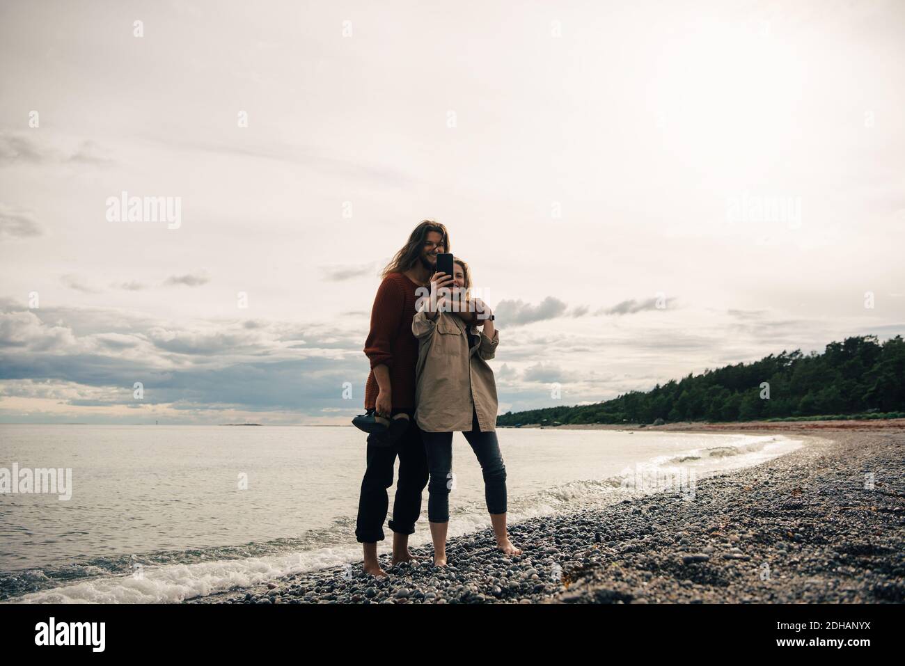 Une longueur totale de couple qui prend son selfie sur son smartphone à plage contre le ciel Banque D'Images