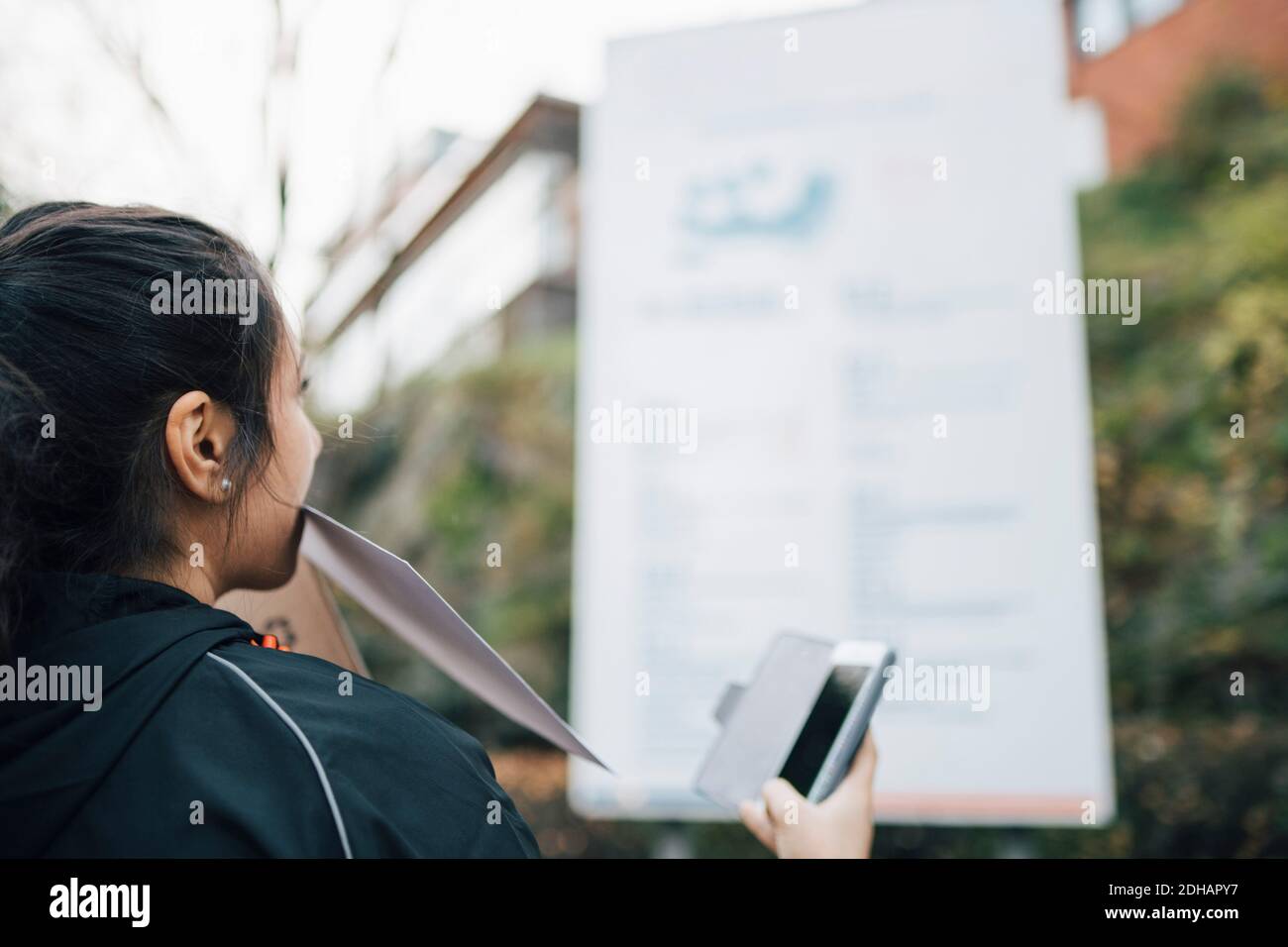 Messager féminin portant un document dans la bouche et tenant un smartphone en regardant le panneau d'affichage Banque D'Images