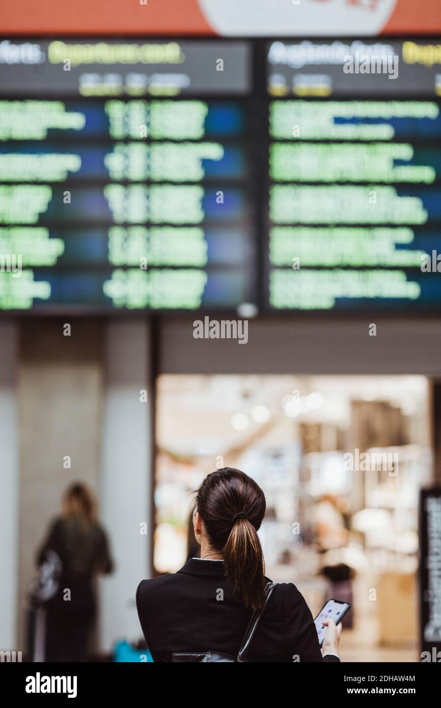 Vue arrière d'une femme d'affaires tenant un smartphone tout en regardant arrivée départ à la gare Banque D'Images