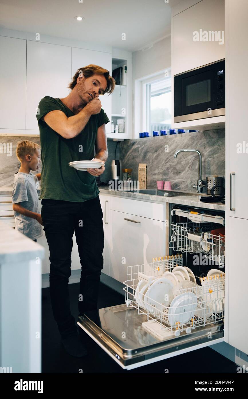 Homme parlant sur un téléphone portable tout en utilisant le lave-vaisselle dans la cuisine Banque D'Images