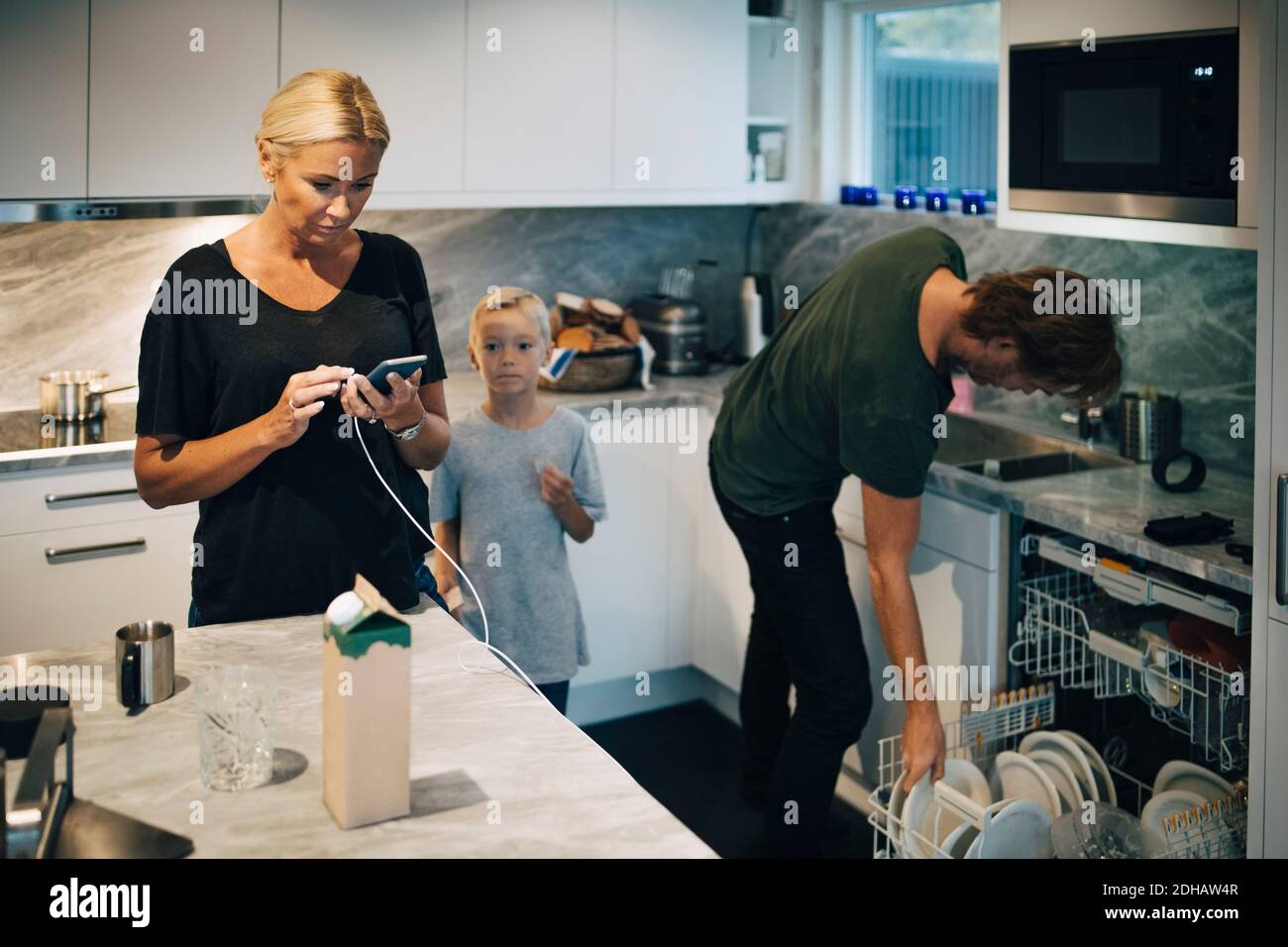 Garçon debout par la mère en utilisant le téléphone portable et le nettoyage de père plaques au lave-vaisselle dans la cuisine Banque D'Images