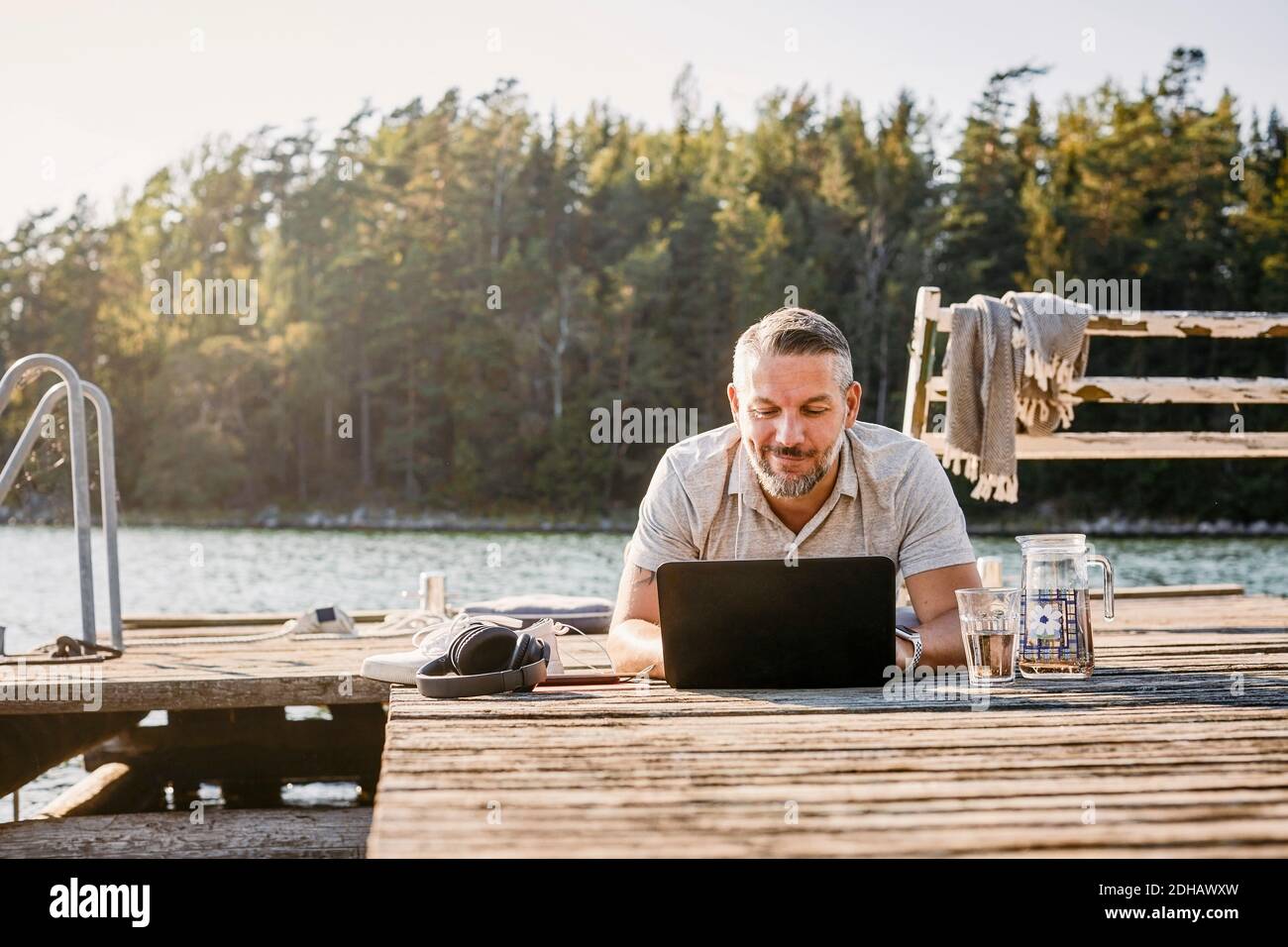 Homme souriant couché sur le devant tout en utilisant un ordinateur portable sur du bois jetée Banque D'Images