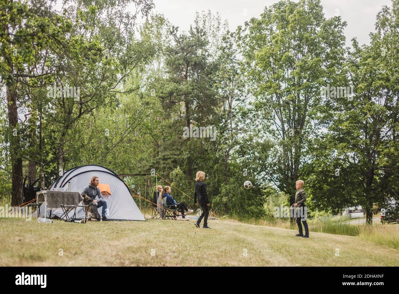 Les enfants jouent au football tandis que la famille s'assoit sous la tente camping Banque D'Images