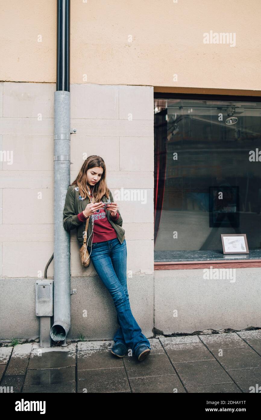 Pleine longueur de fille utilisant un smartphone tout en s'appuyant sur tuyau contre le mur Banque D'Images