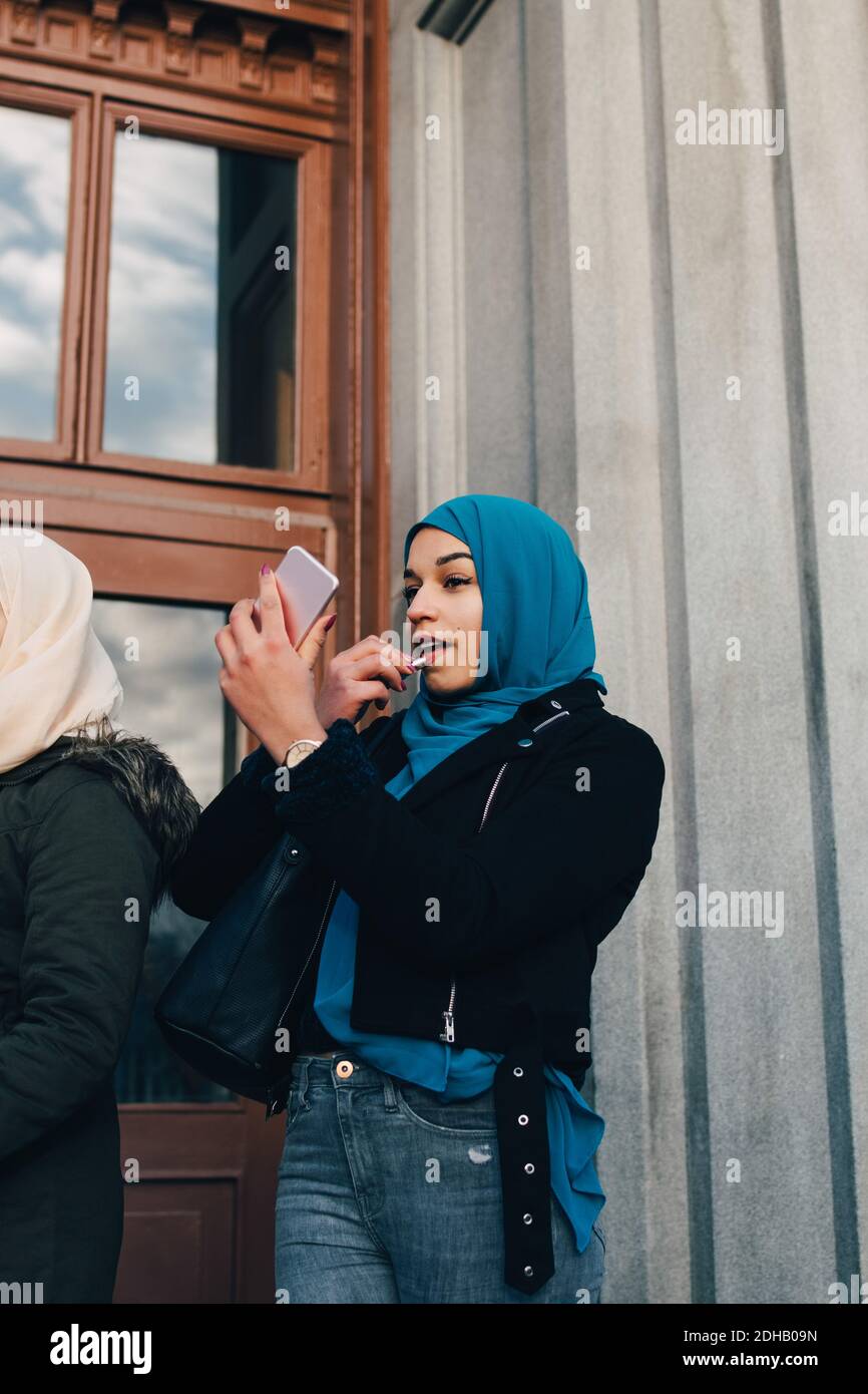 Vue à angle bas de la jeune femme musulmane appliquant le regard de rouge à lèvres dans le téléphone mobile par un ami contre la construction Banque D'Images