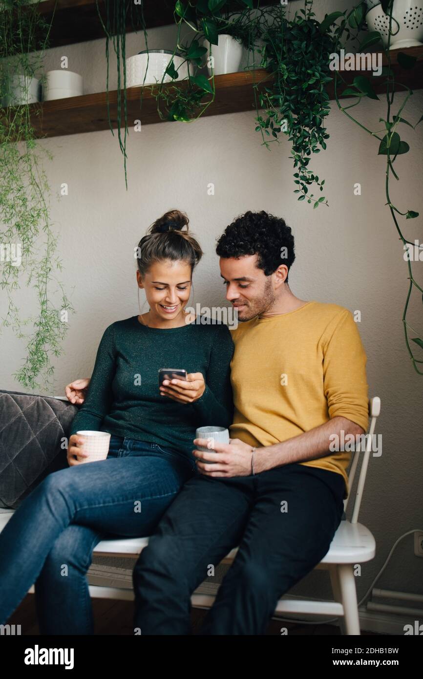 Petit ami souriant et petite amie regardant le téléphone mobile tout en ayant café dans la salle de séjour Banque D'Images