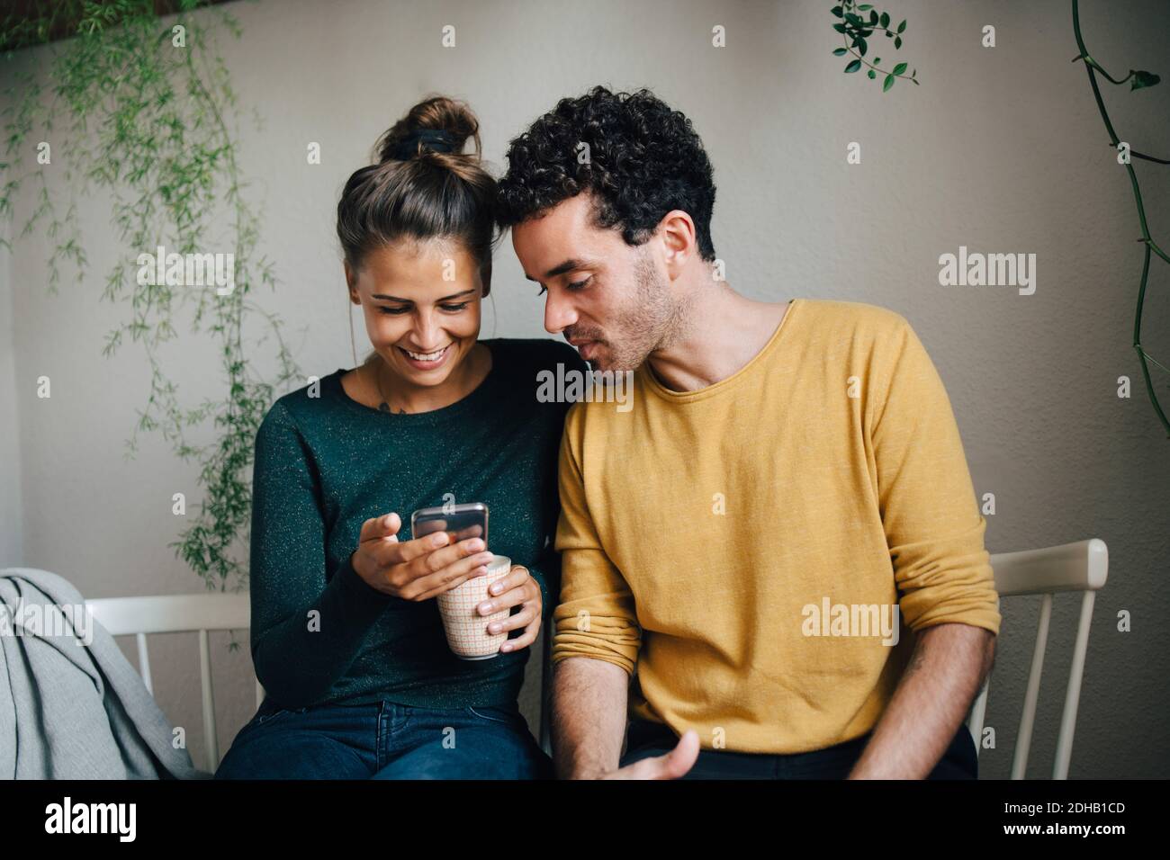 Petite amie souriante montrant un smartphone à un ami tout en prenant un café dans la salle de séjour Banque D'Images