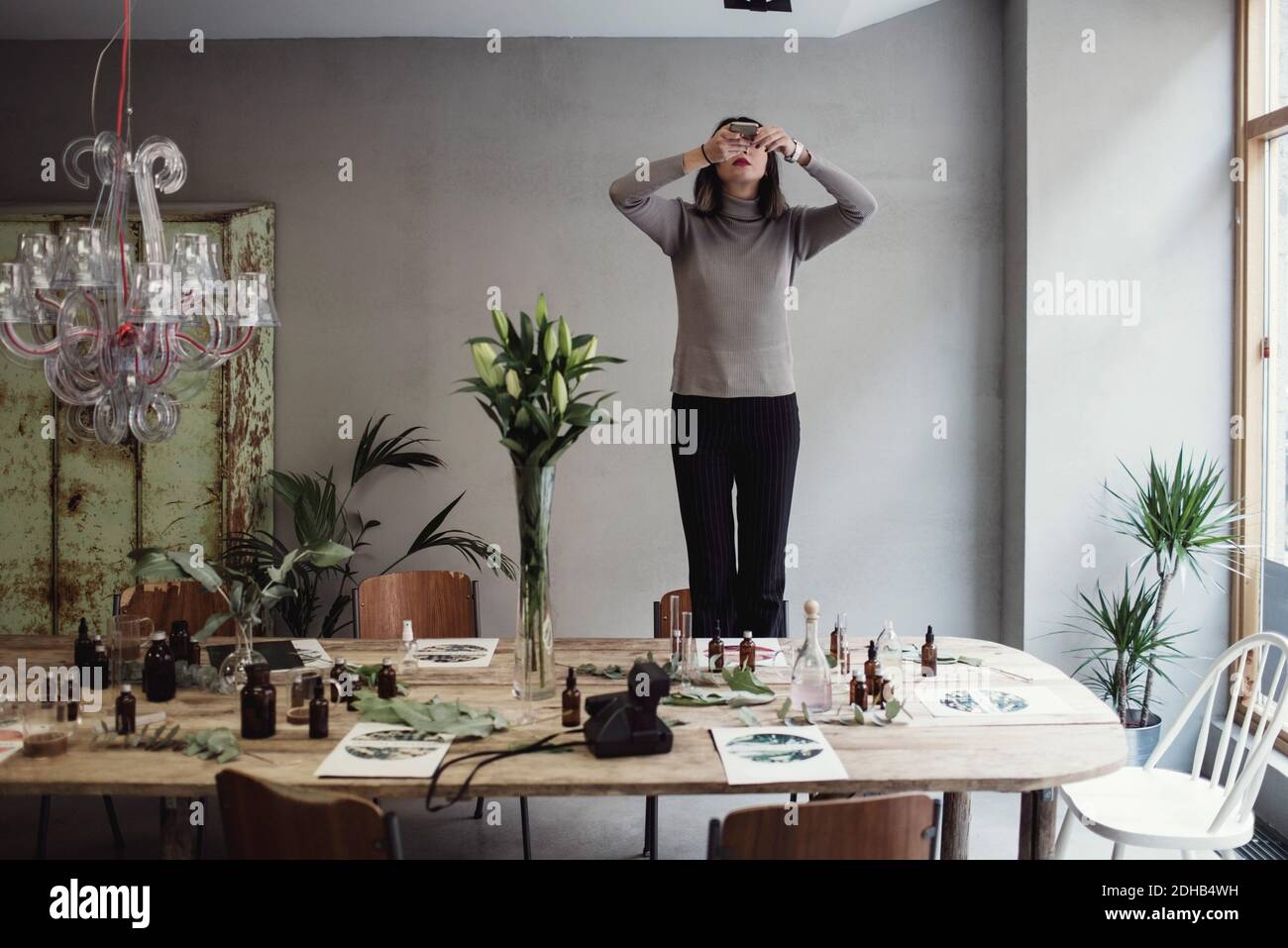 Jeune femme debout sur une chaise, photographiant des flacons de parfum sur une table en atelier Banque D'Images