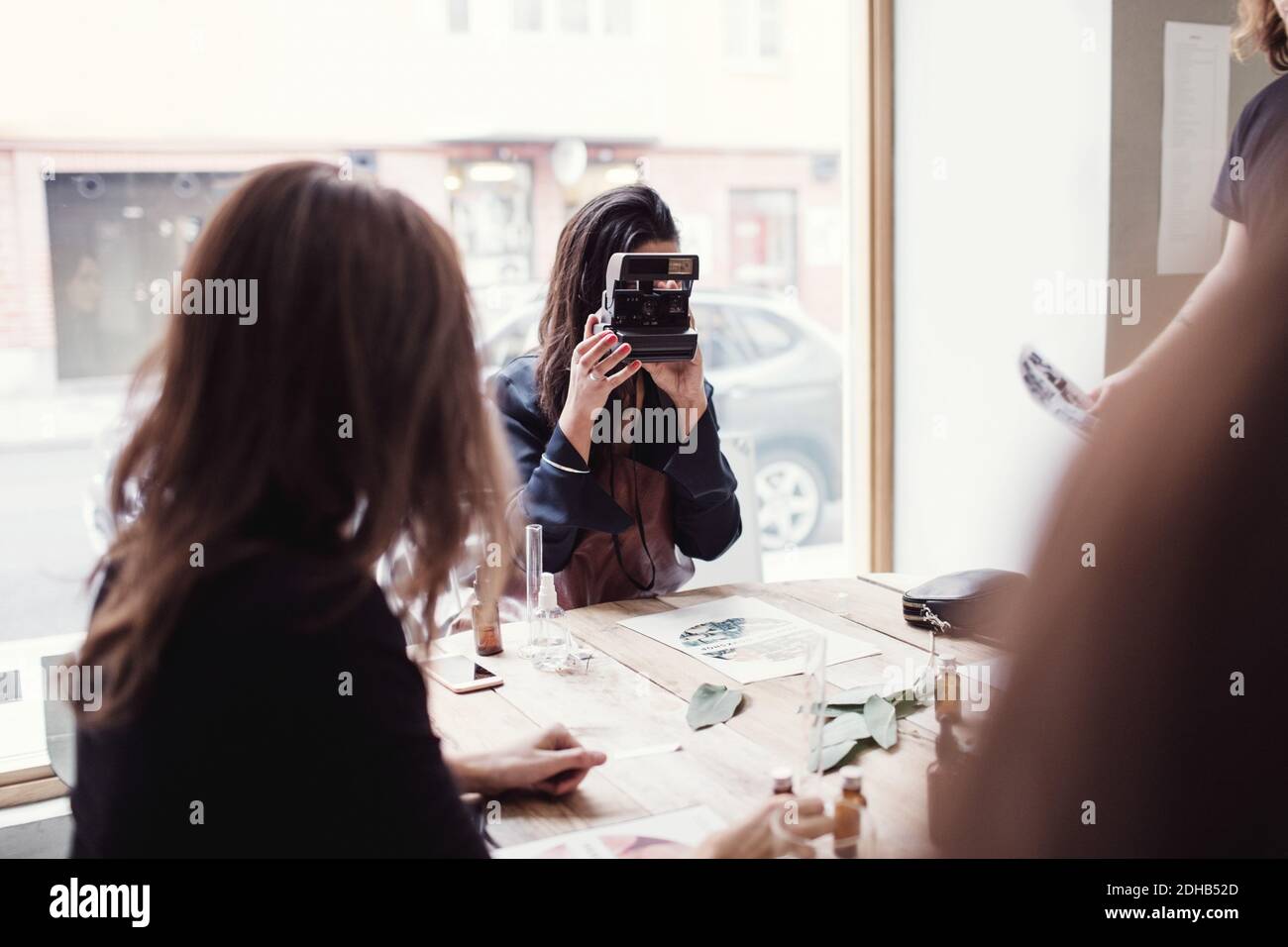 Femme photographiant en étant assise par un collègue à la table contre la fenêtre en atelier Banque D'Images