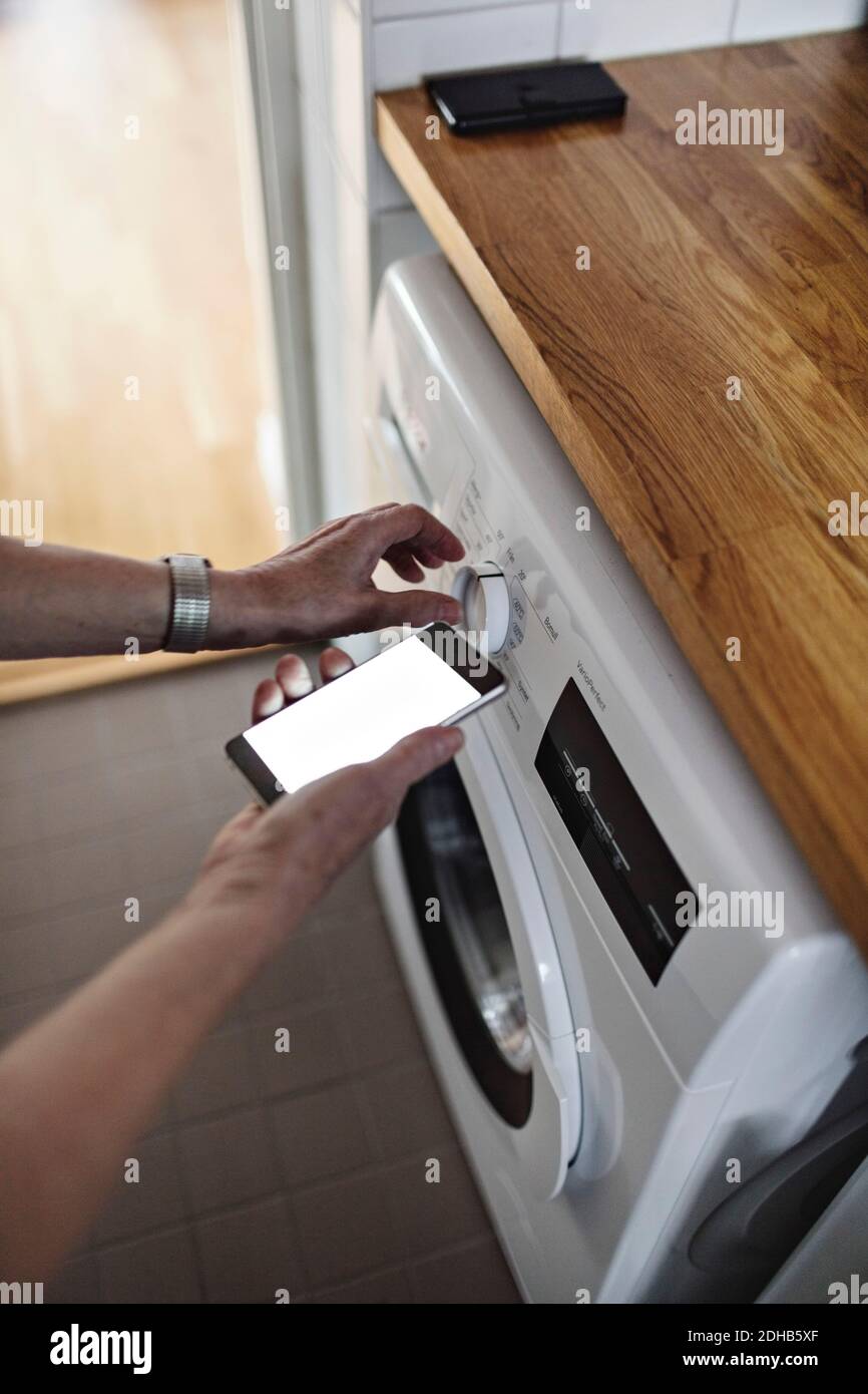 Les mains d'une femme âgée qui tient un téléphone portable pendant son utilisation sont coupées machine à laver dans la salle de bains à la maison Banque D'Images