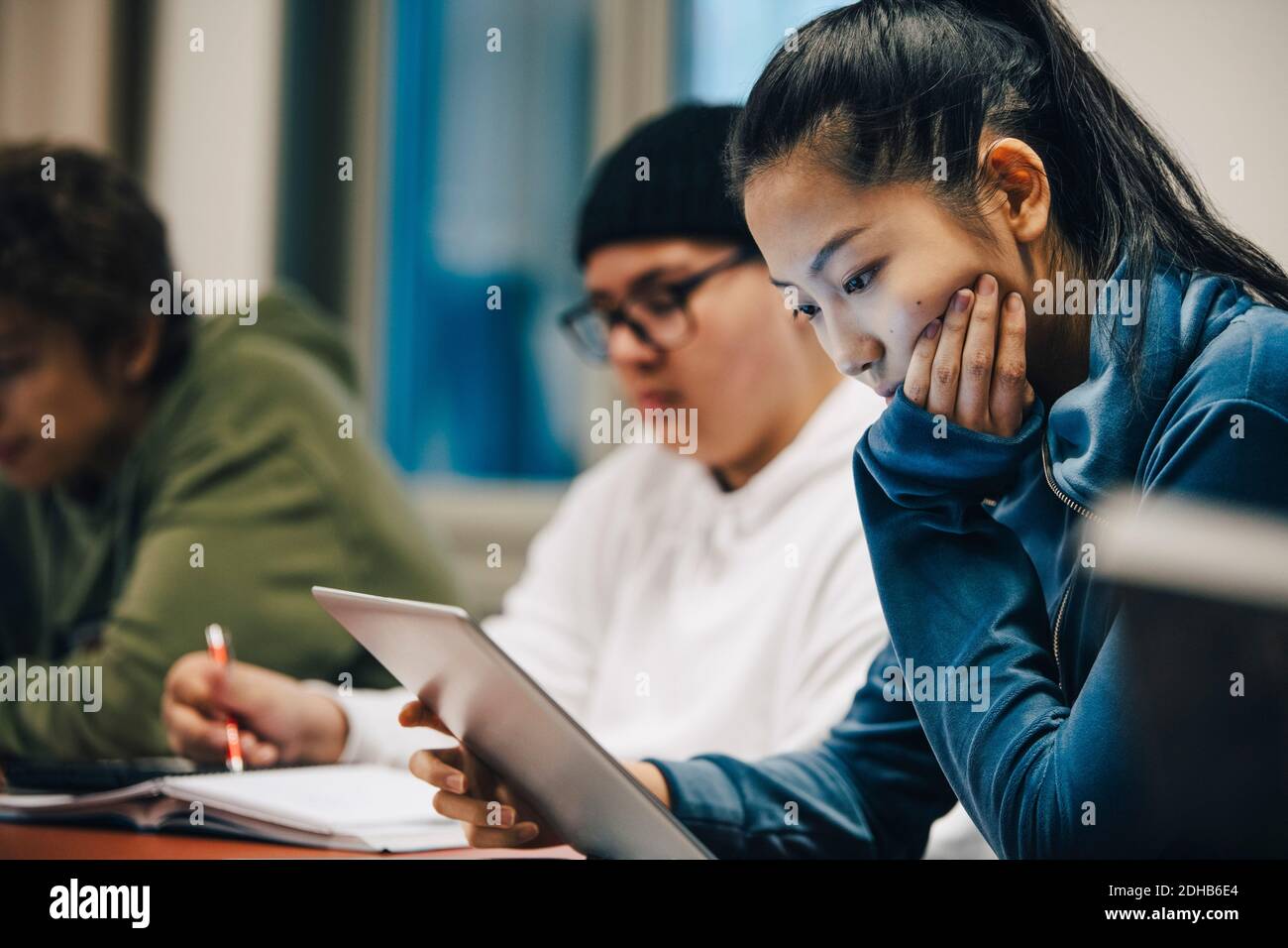 Adolescente avec la main sur le menton en utilisant la tablette numérique pendant assis par des camarades de classe à l'école secondaire Banque D'Images