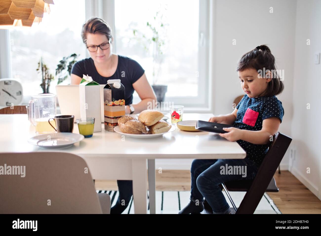 Mère utilisant un ordinateur portable tout en étant assise avec sa fille pendant le petit déjeuner à tableau Banque D'Images