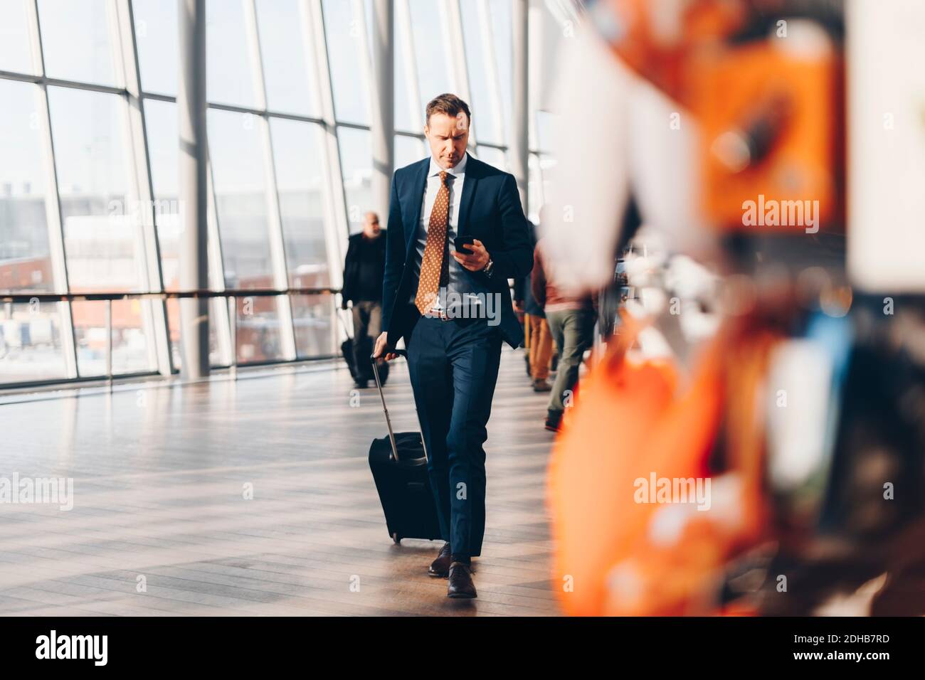 Pleine longueur d'homme d'affaires mature avec bagages à l'aide d'un téléphone portable en marchant dans le terminal de l'aéroport Banque D'Images