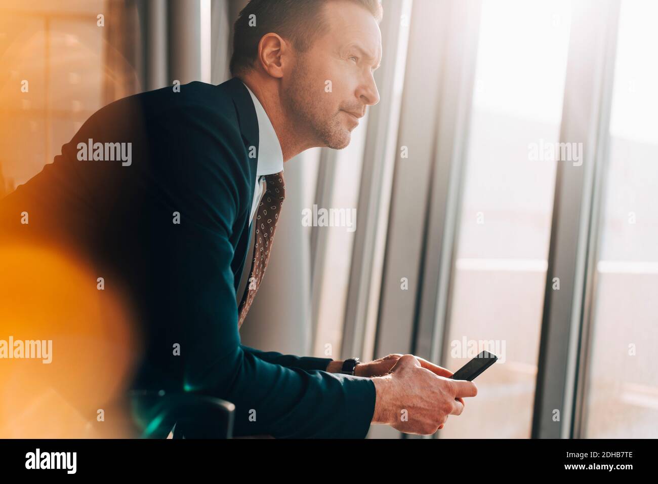 Vue latérale d'un homme d'affaires mature tenant un téléphone portable tout en regardant par la fenêtre de l'aéroport Banque D'Images